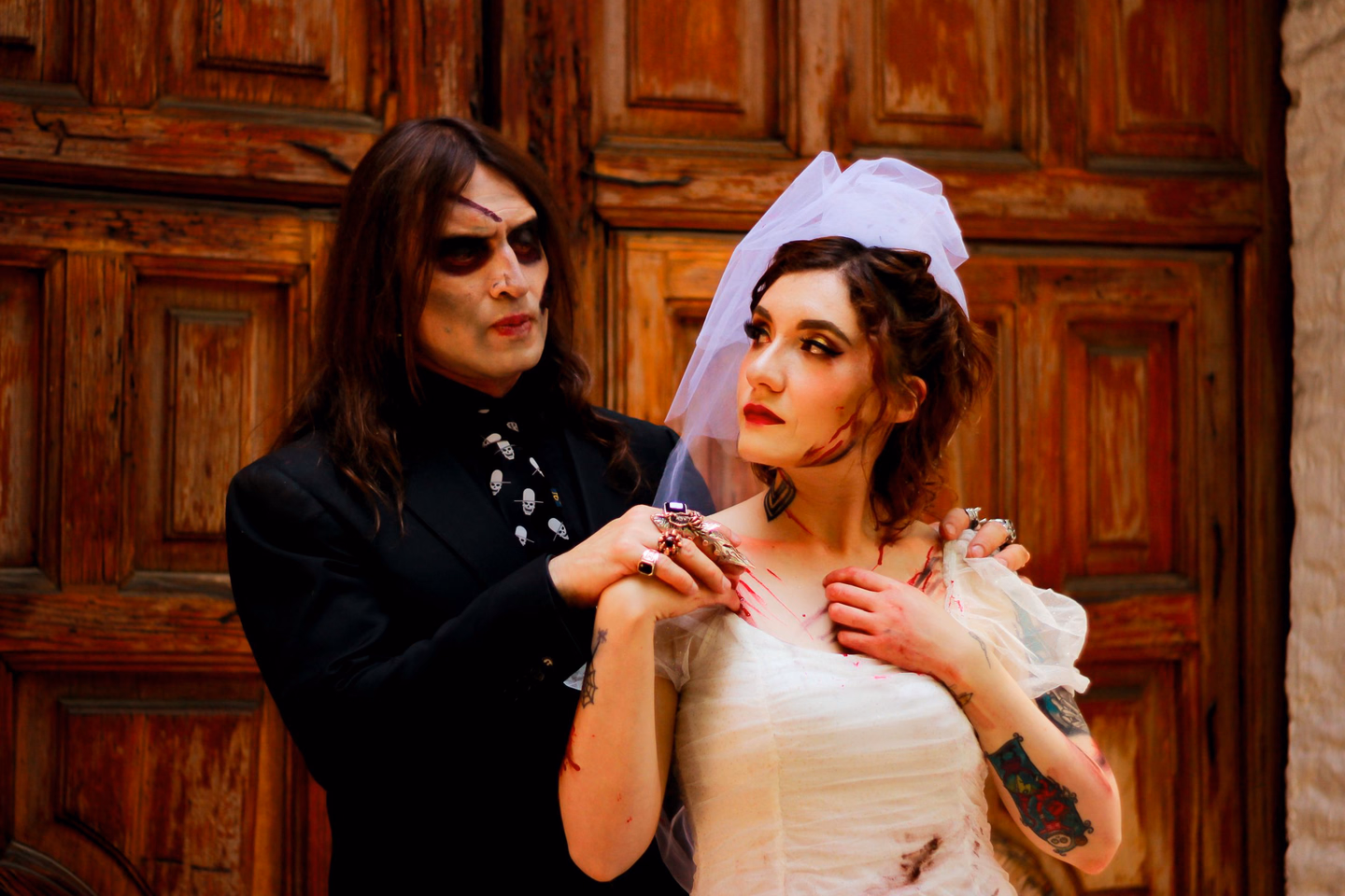 A man in a black suit and skull-print tie with dark eye makeup standing behind a woman in a torn white wedding dress and short veil, both with stage blood on their hands, in front of a carved wooden door