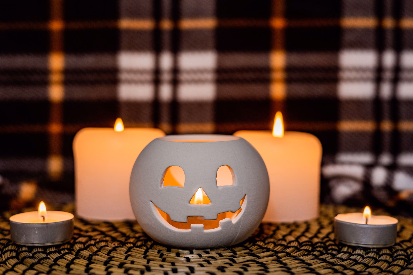 A white ceramic jack-o'-lantern between two lit pillar candles and two tea lights on a woven mat