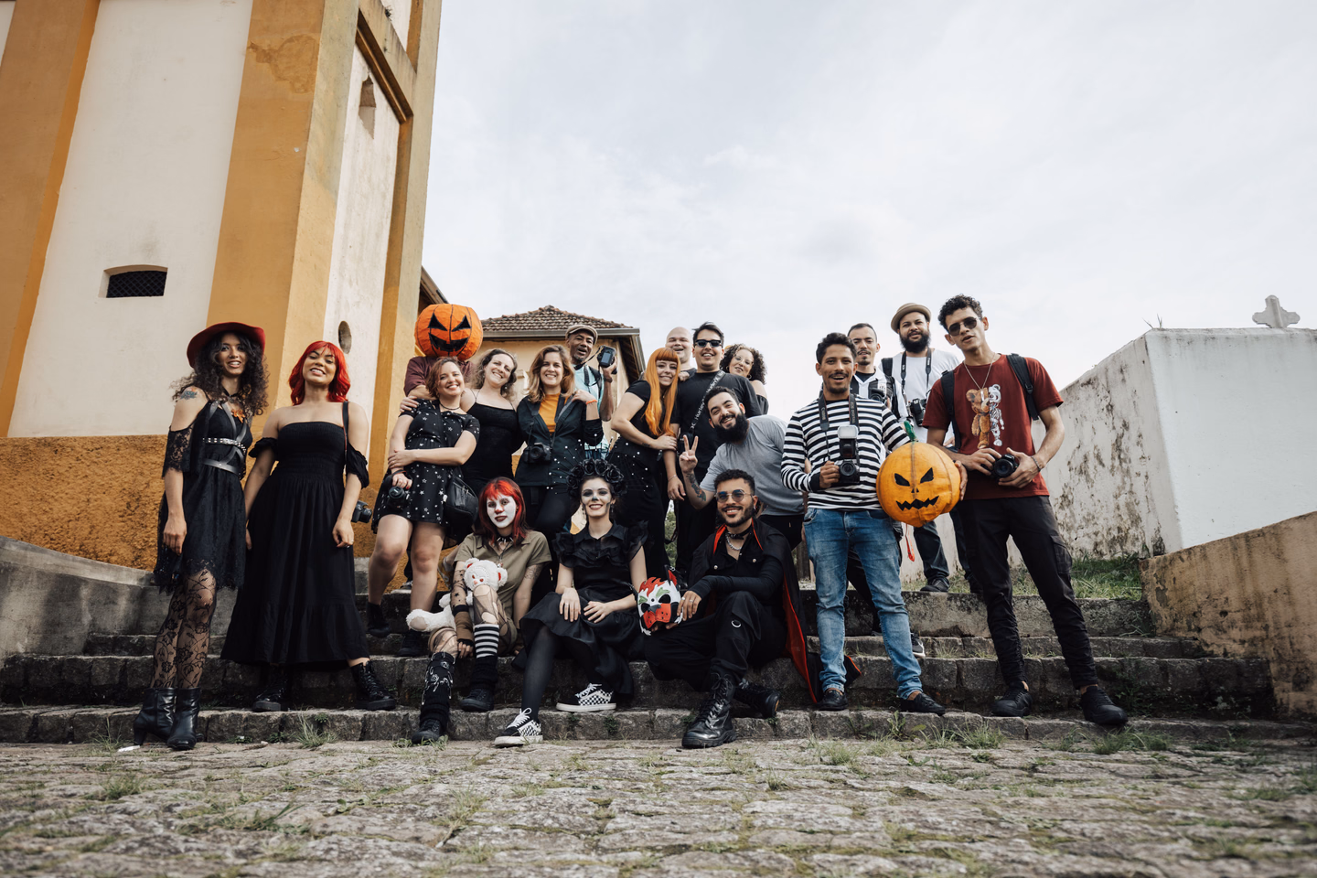 A large group of adults on old stone steps beside a yellow-trimmed building, some in a witch hat, black dresses, clown makeup and a pumpkin-head mask, others in jeans and T-shirts holding cameras and pumpkin props