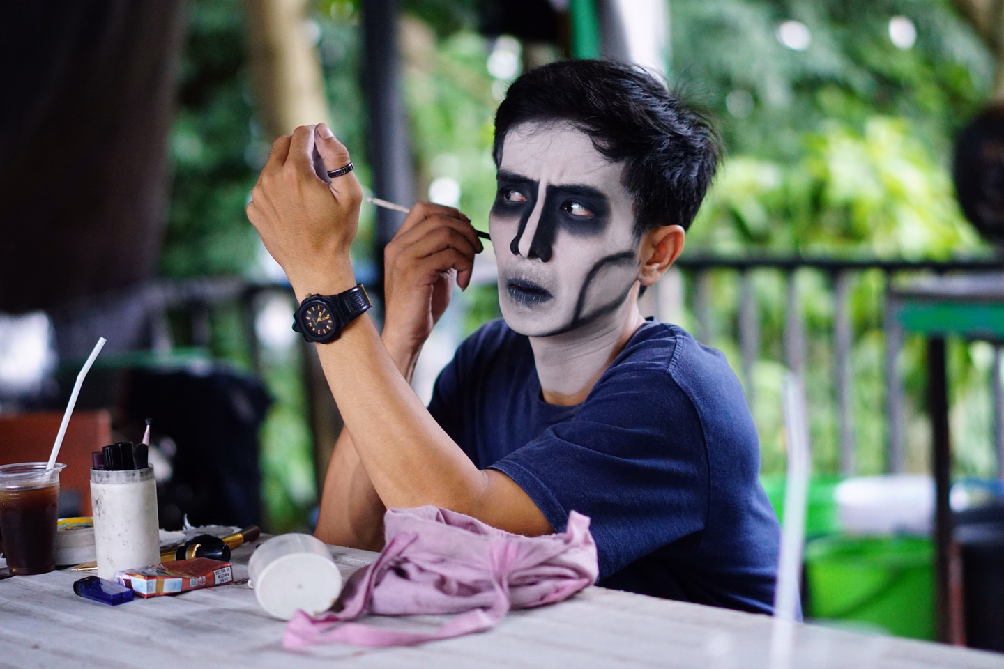 A man in a navy T-shirt painting black shapes over white skull face paint at an outdoor table covered with makeup pots and brushes