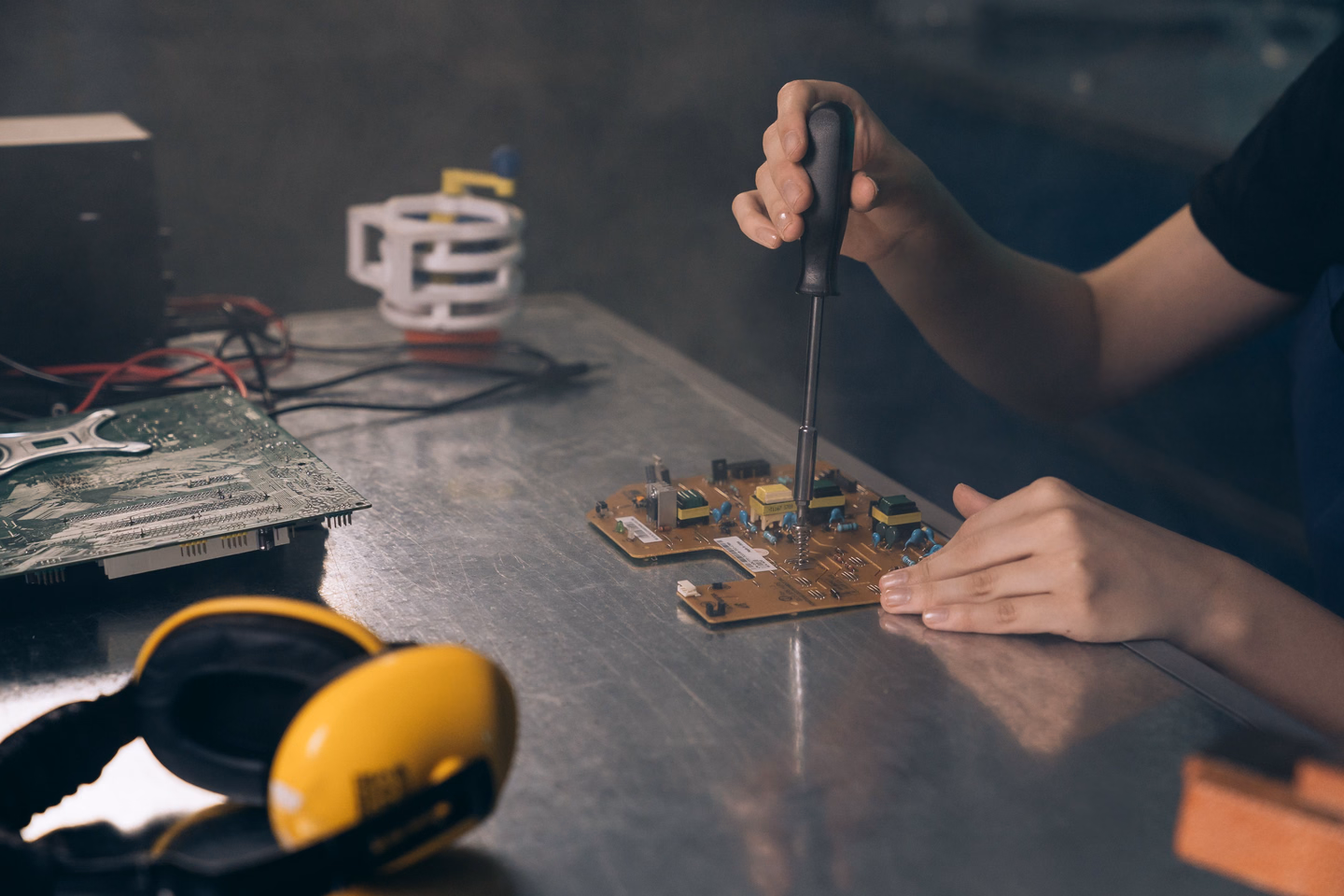 Hands using a small screwdriver on a green circuit board on a dark workbench, with a pair of yellow ear defenders in front