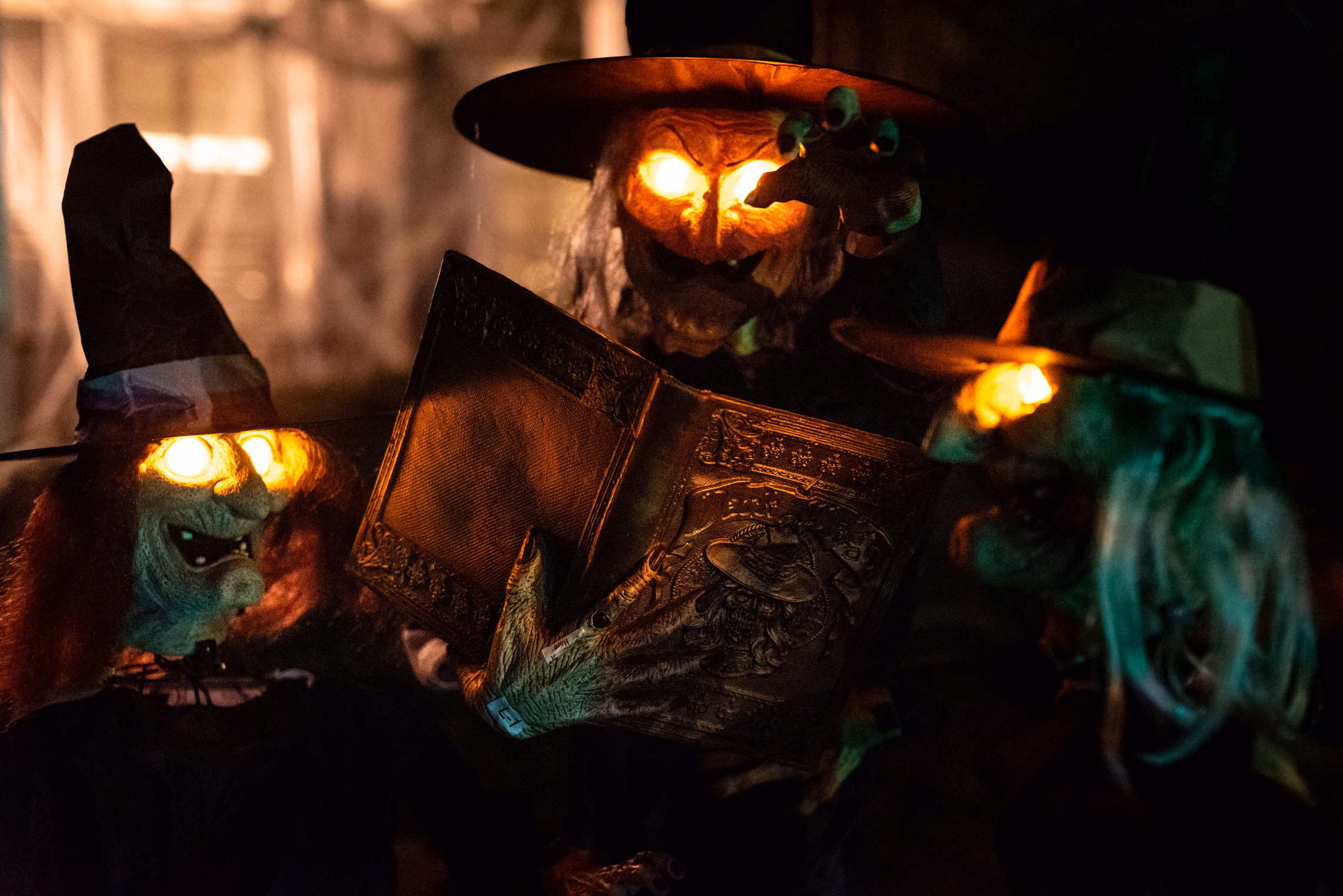 Three witch figures at night with glowing orange eyes, one holding an open spell book, against a dark background