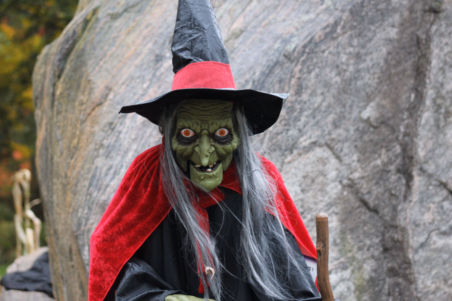 A witch figure with a green face, orange eyes and long gray hair under a black and red pointed hat, standing outdoors against a large rock
