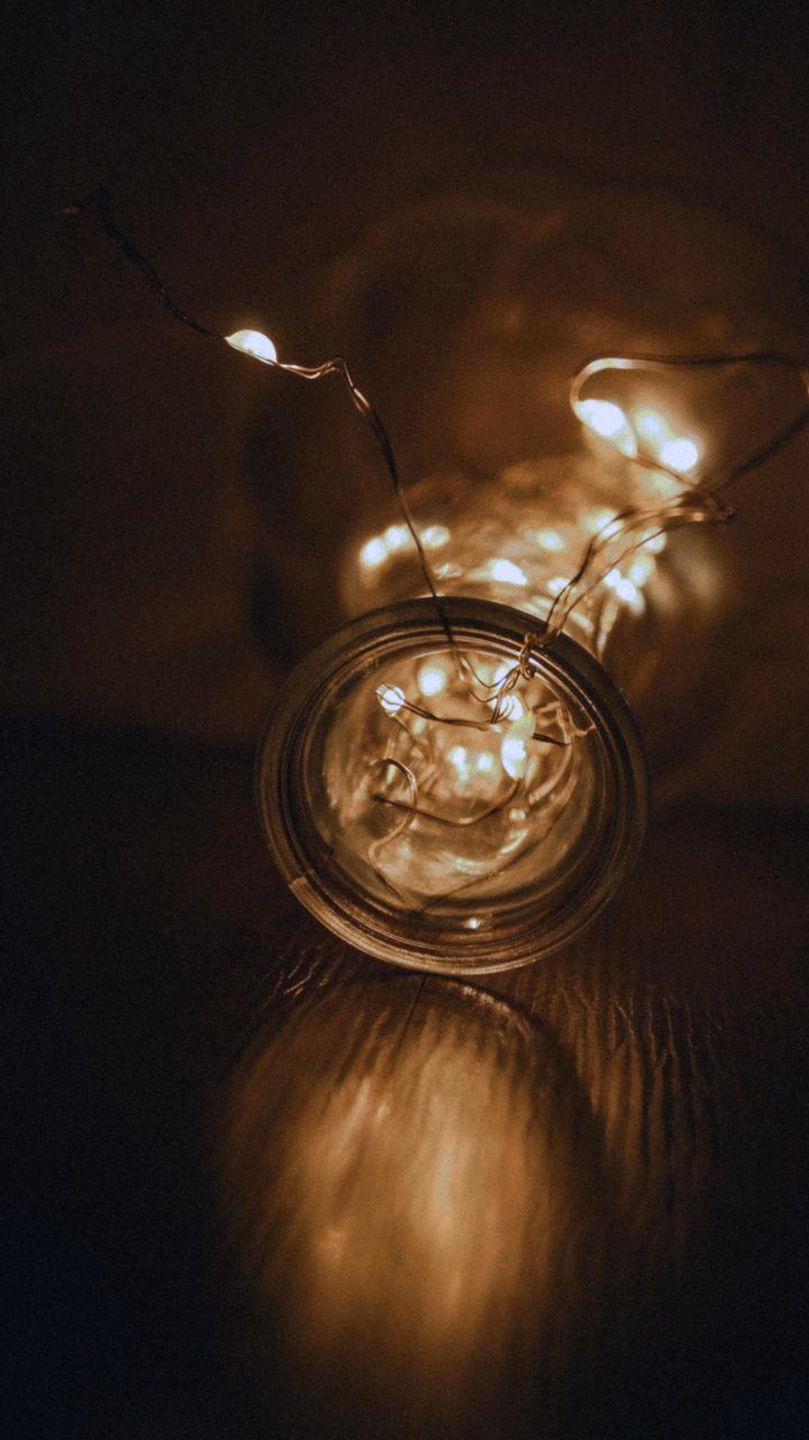 A clear glass bottle on its side glowing with a tangle of warm white string lights inside, against a dark background