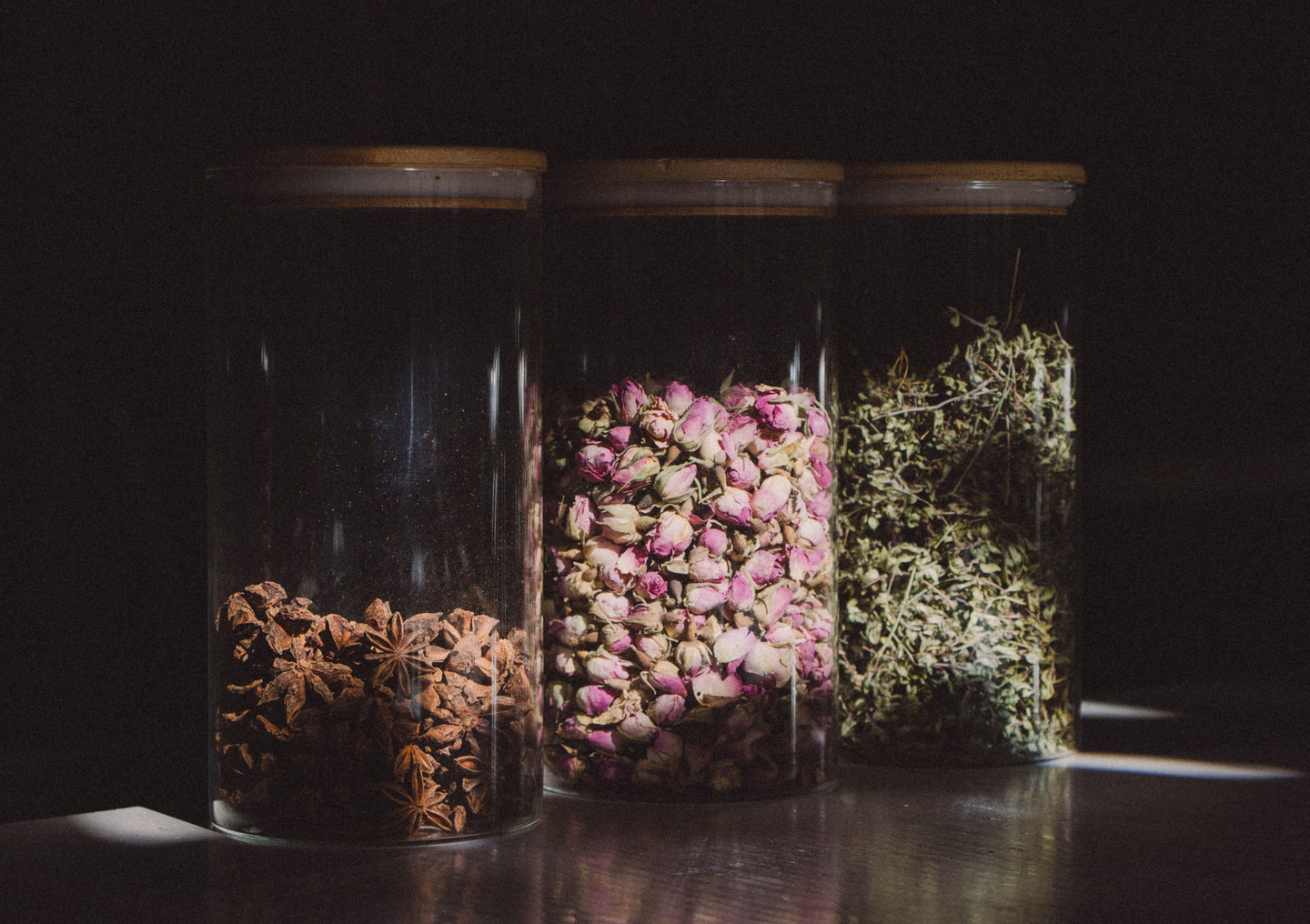 Three tall glass jars in low light, filled with star anise, dried rosebuds and dried herbs