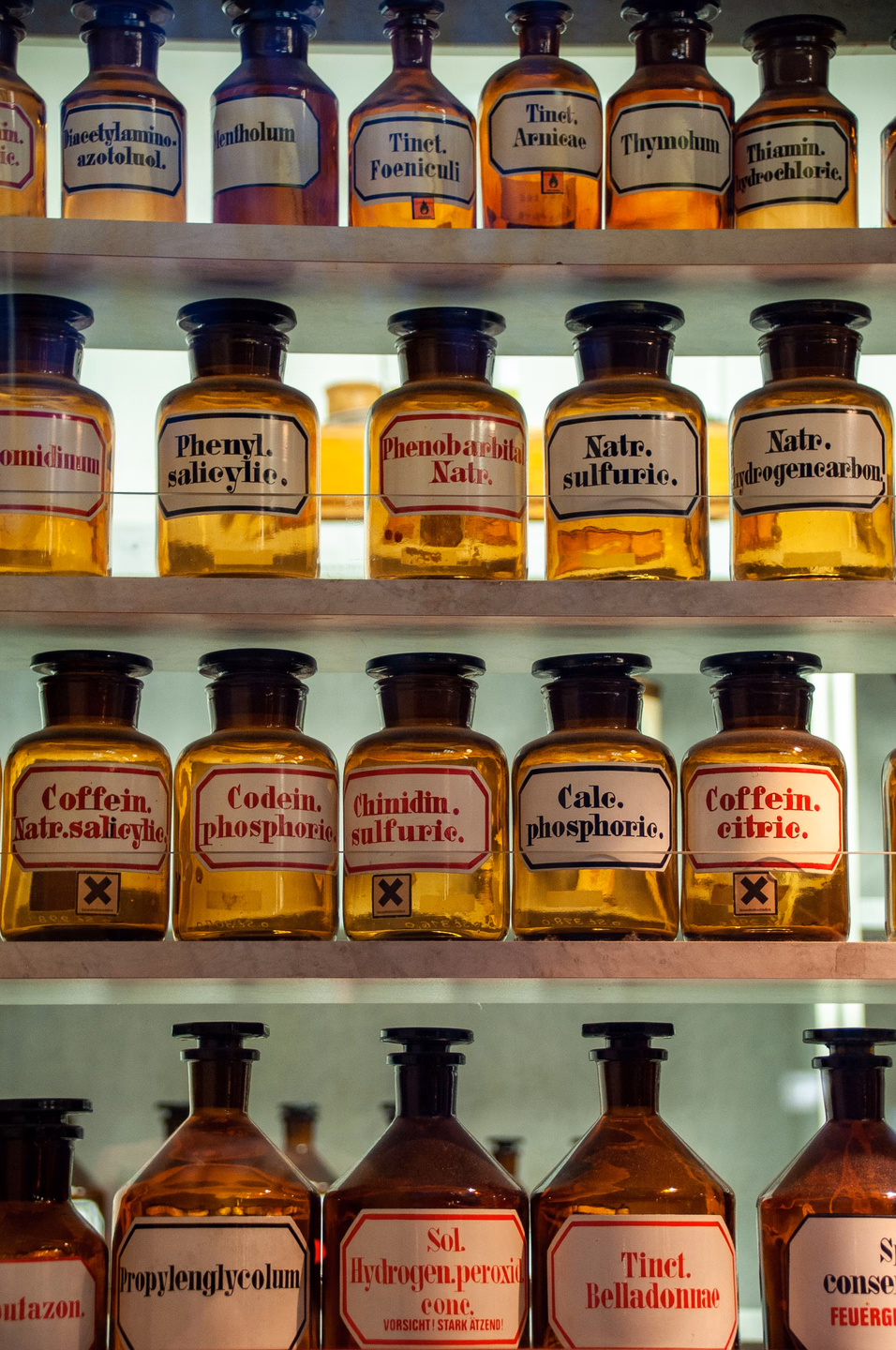 Four shelves of identical amber glass pharmacy bottles with matching white labels outlined in black or red, reading Tinct. Arnicae, Tinct. Belladonnae and Codein. phosphoric.