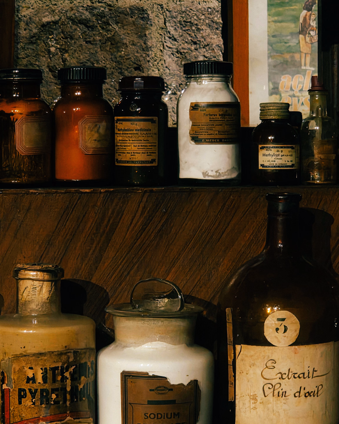A mismatched collection of old brown, amber and white pharmacy jars on a wooden shelf, one large bottle with a handwritten label reading Extrait Clin d'oeil