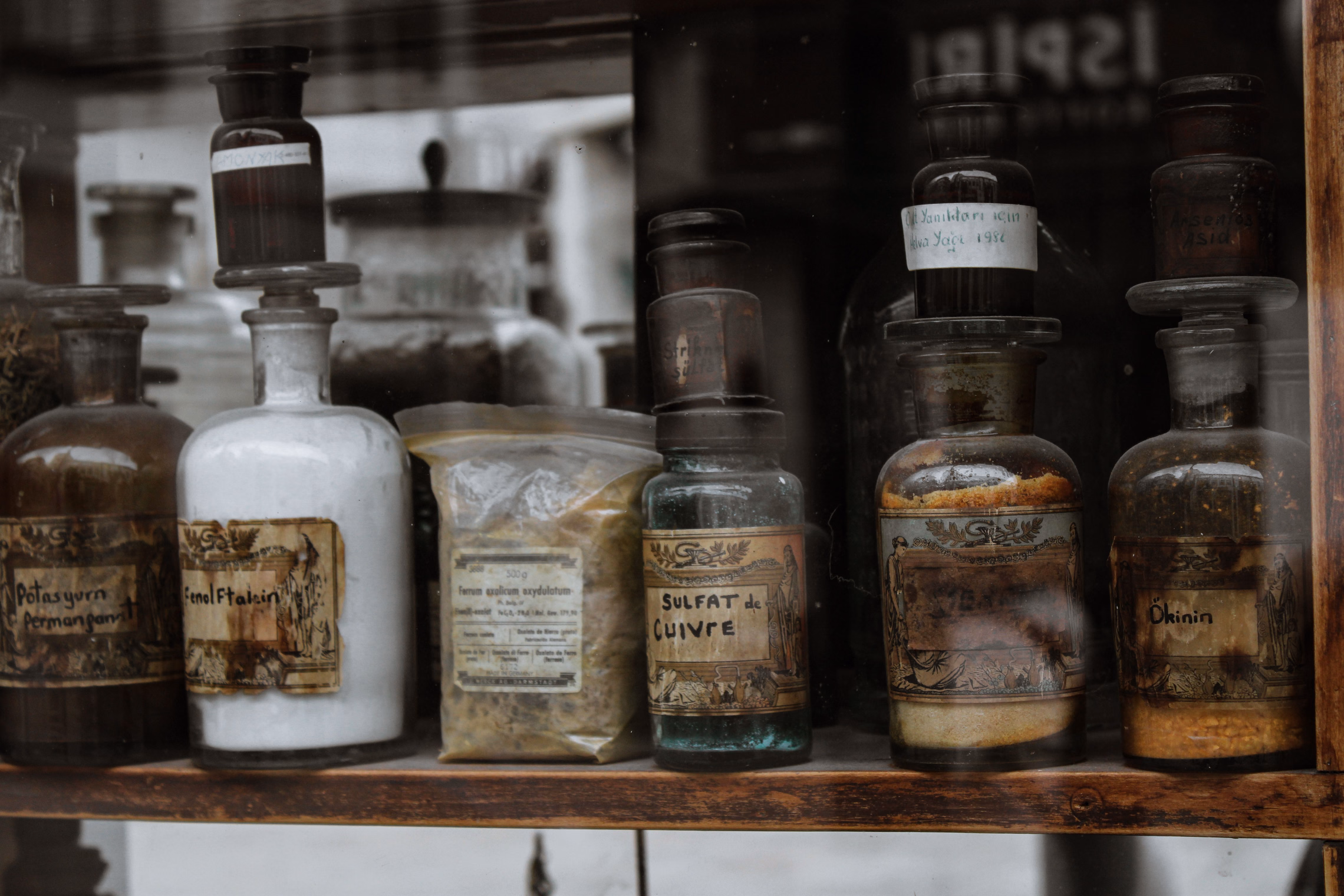 Old glass apothecary bottles on a wooden shelf behind a glass pane, with faded, ornate paper labels reading Fenol Ftalein and Sulfat de Cuivre