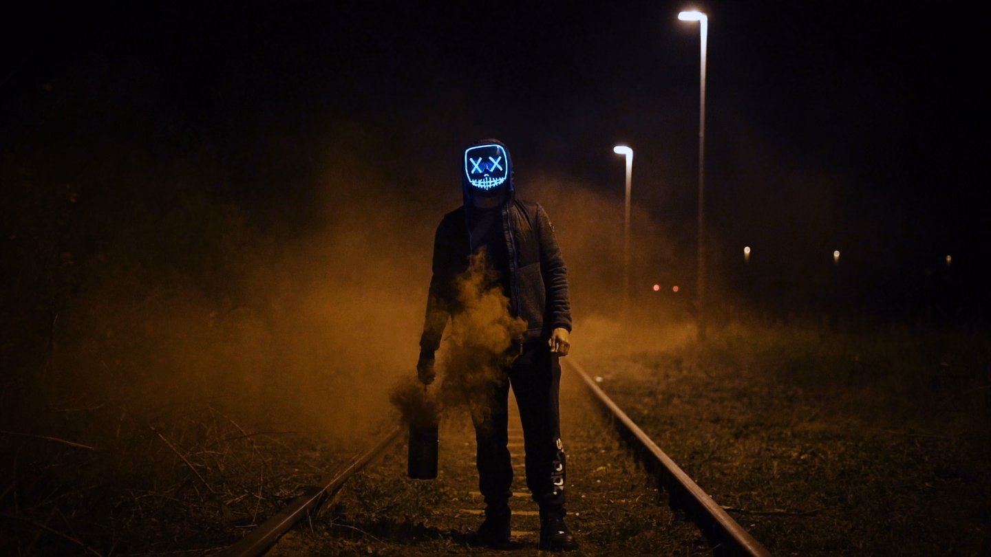 A figure in a glowing LED mask standing on railroad tracks at night, wrapped in low mist