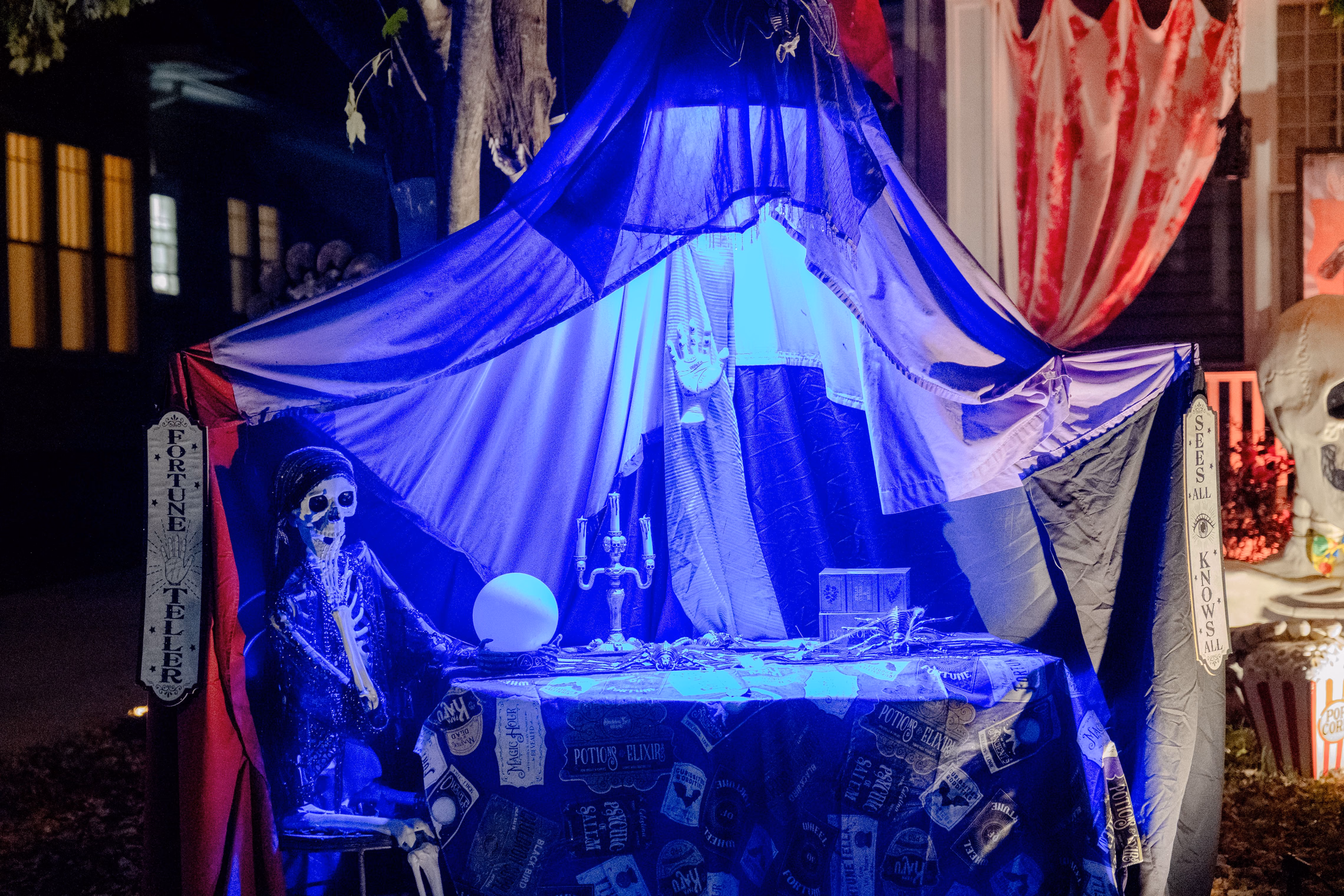 A fortune teller booth outside a house at night, with skeletons in the chairs lit purple and blue