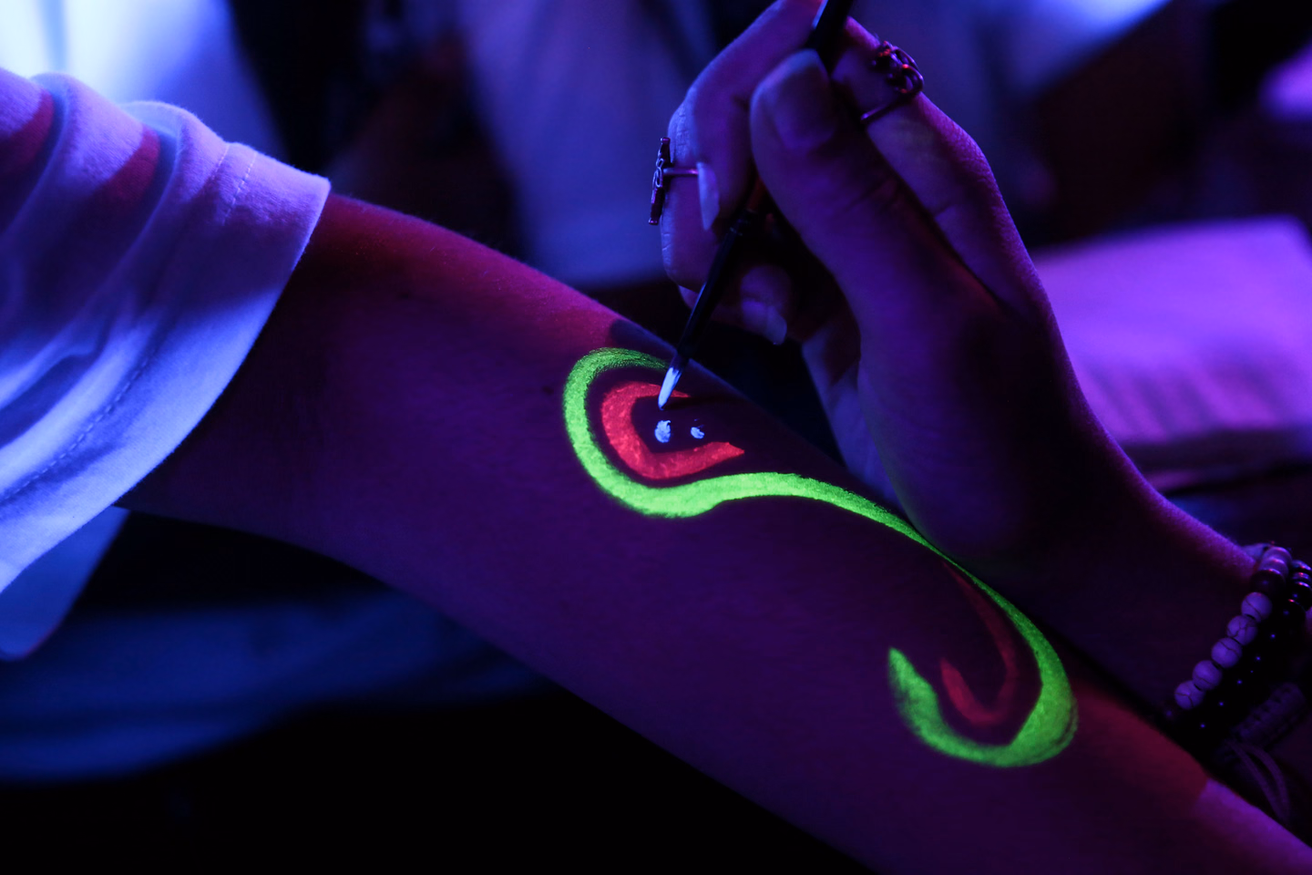 A hand holding a fine brush paints a glowing green and red swirl onto a forearm under blacklight