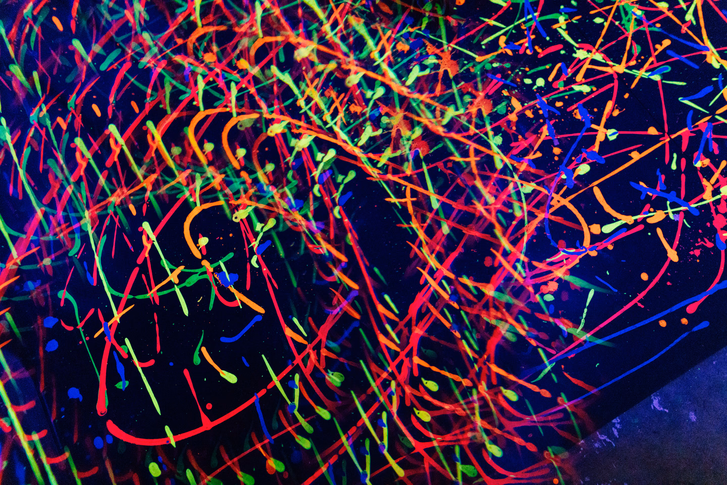 Splattered lines of neon green, orange, pink and blue paint glowing across a black surface under ultraviolet light