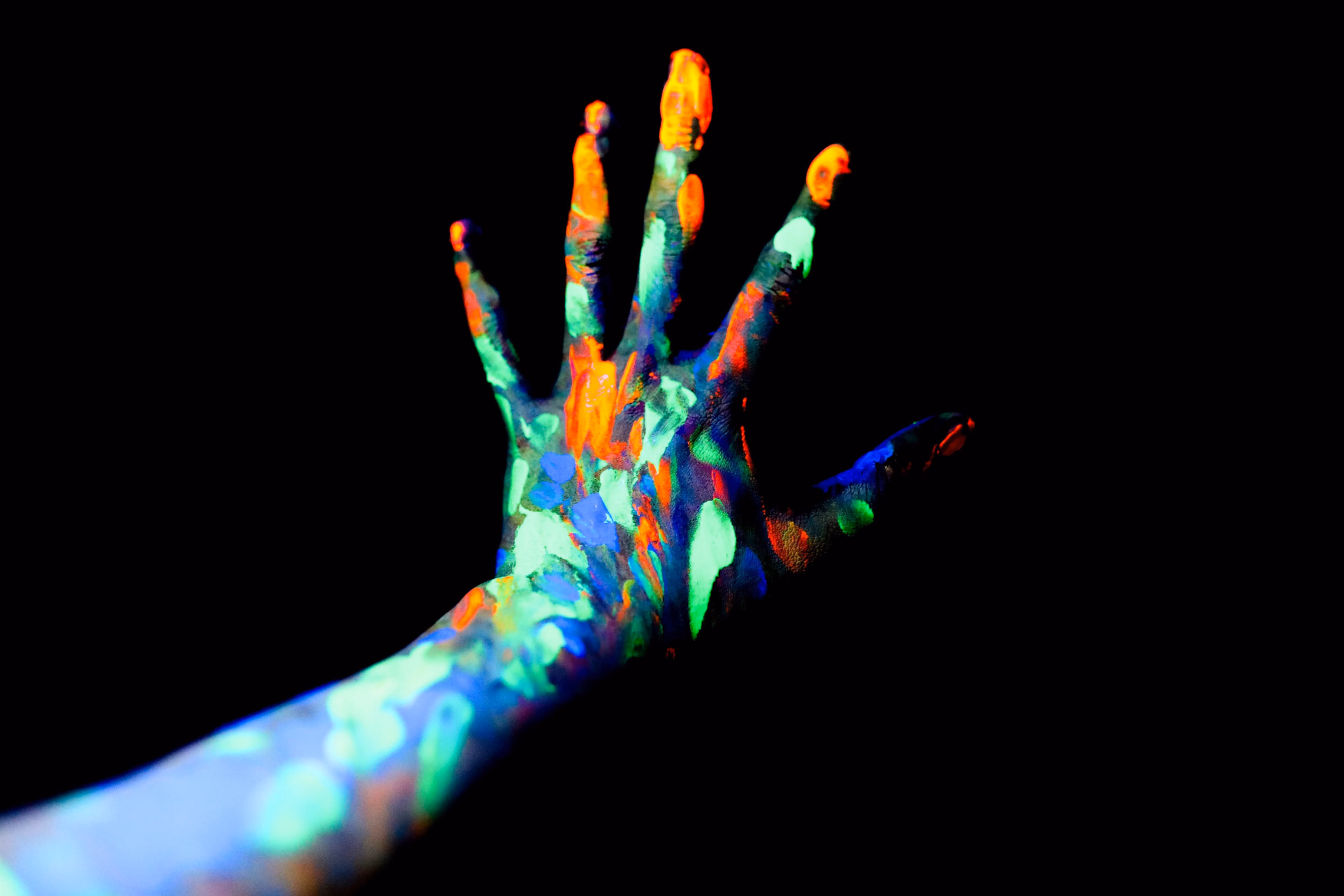 An outstretched hand and forearm smeared with green, orange and blue fluorescent paint, glowing against total darkness under blacklight