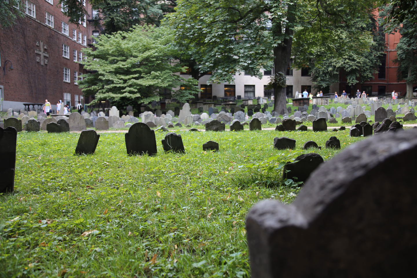 Short, dark slate headstones scattered across the grass of a historic Boston burying ground, with brick buildings behind