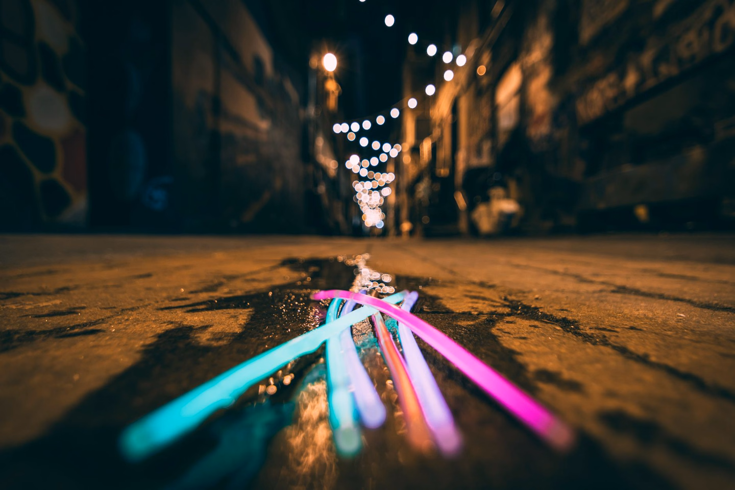 A handful of lit glow sticks in pink, blue and green lying on a wet street at night