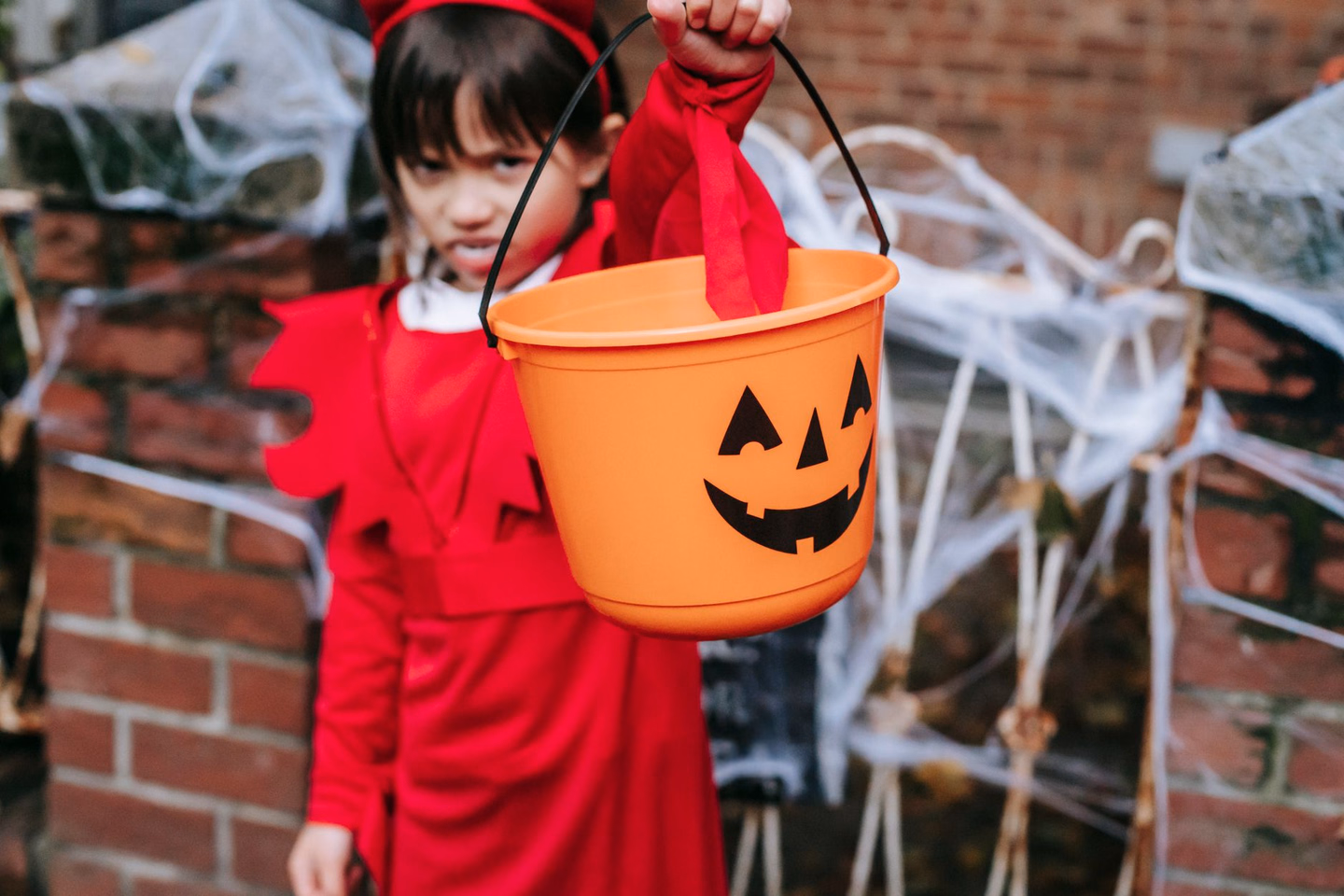 A child in a red costume holding out an orange jack-o'-lantern bucket in front of a brick wall draped with fake cobwebs