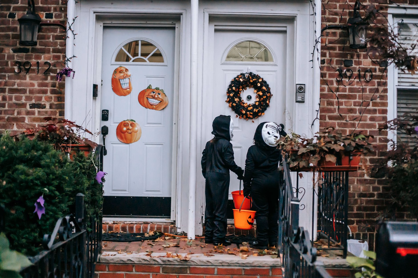 Two children in black skeleton costumes carrying orange pumpkin buckets at a front door decorated with paper jack-o'-lanterns and a wreath
