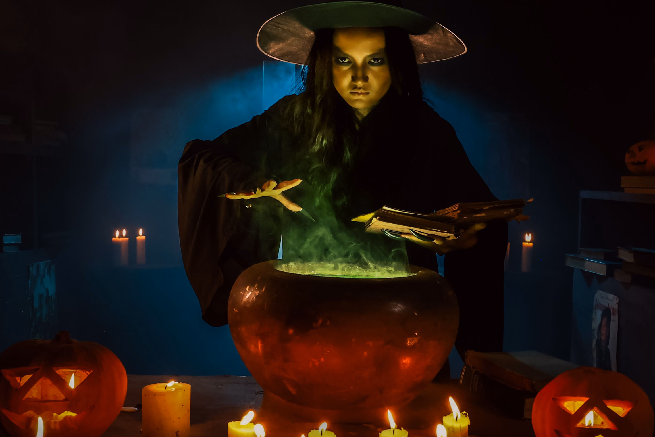 A witch in a wide-brimmed hat bends over a round cauldron glowing green with rising vapor, surrounded by lit candles and two carved jack-o'-lanterns
