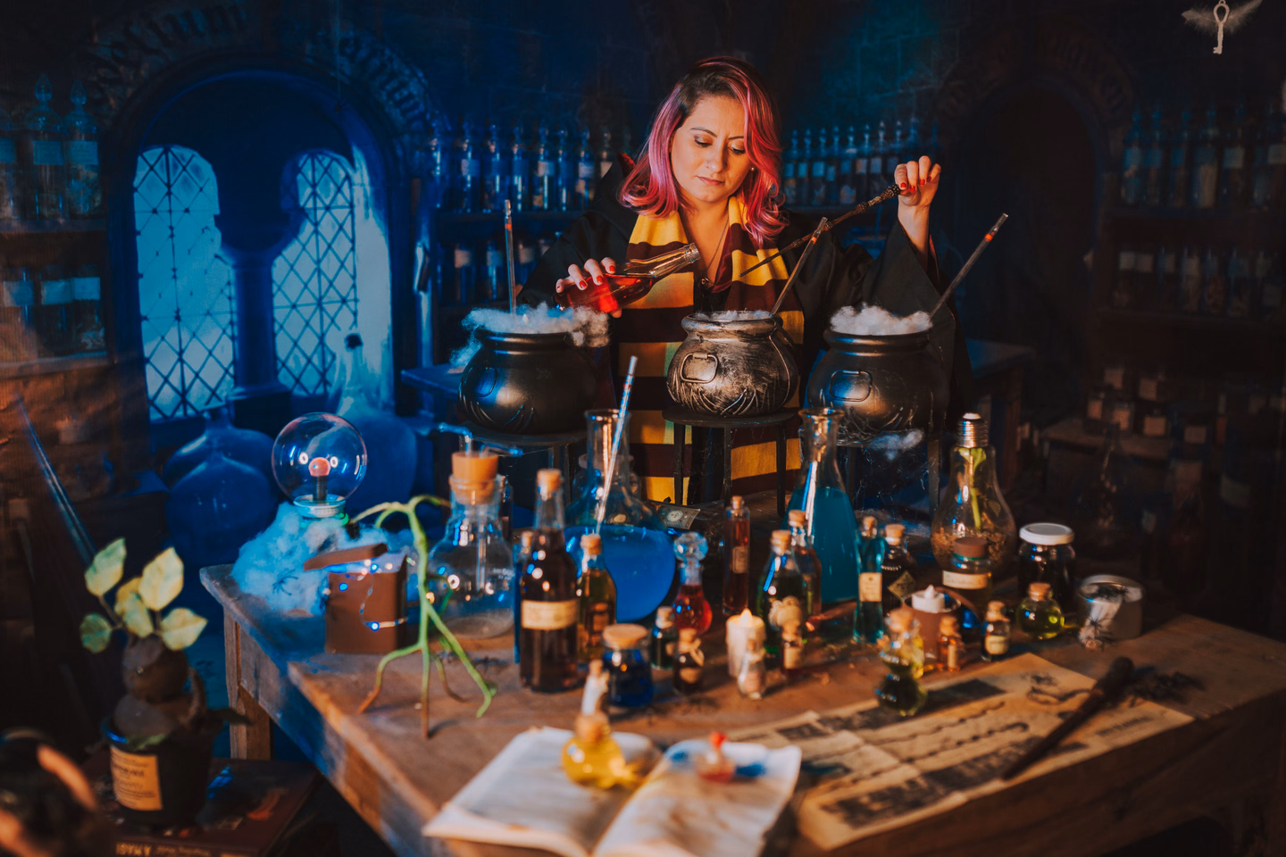 A woman in a witch costume at a crowded potion table covered in cauldrons, glass bottles and open books, lit blue from behind