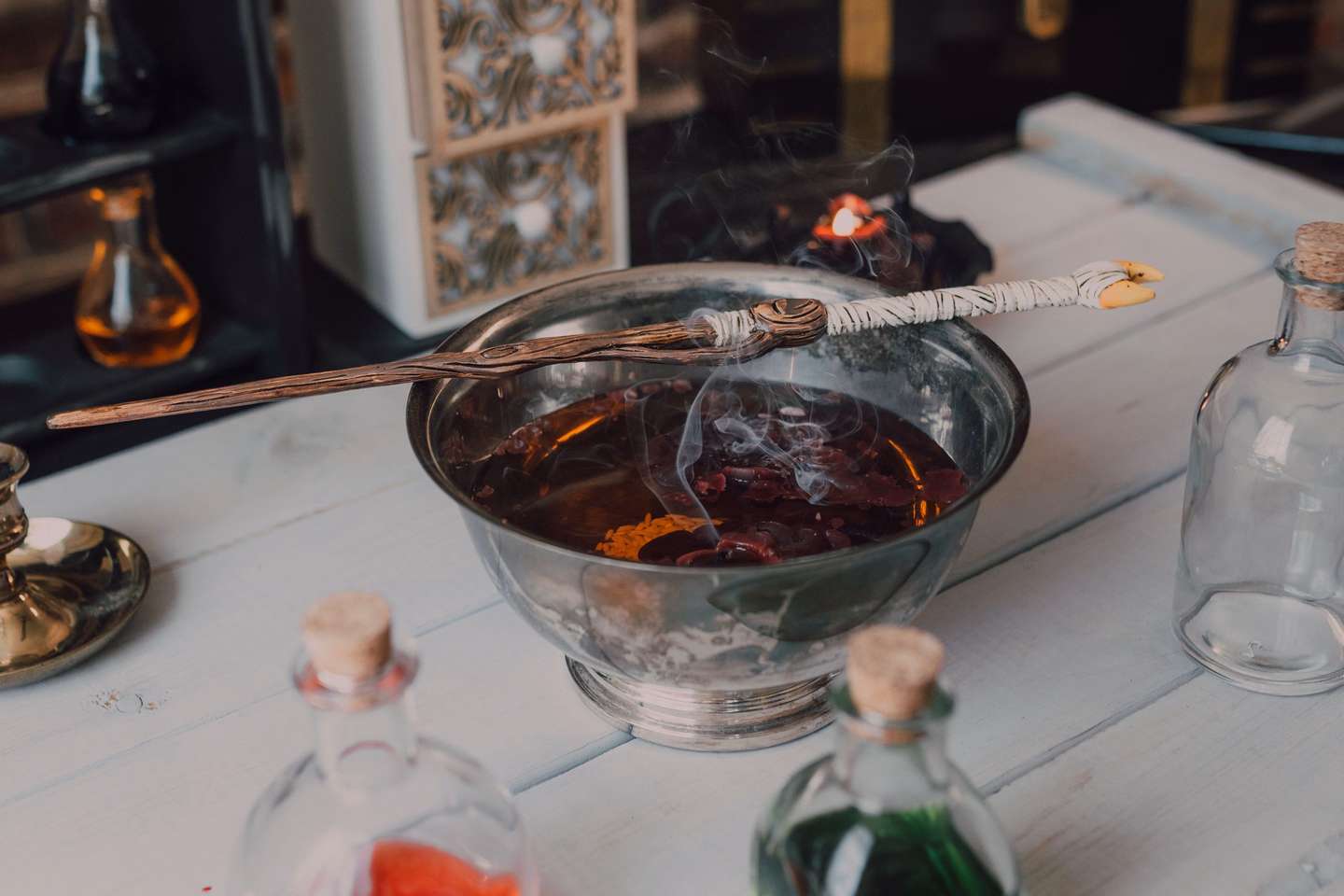 A footed silver bowl of dark red liquid with a wooden wand resting across the rim, surrounded by corked potion bottles on a white table