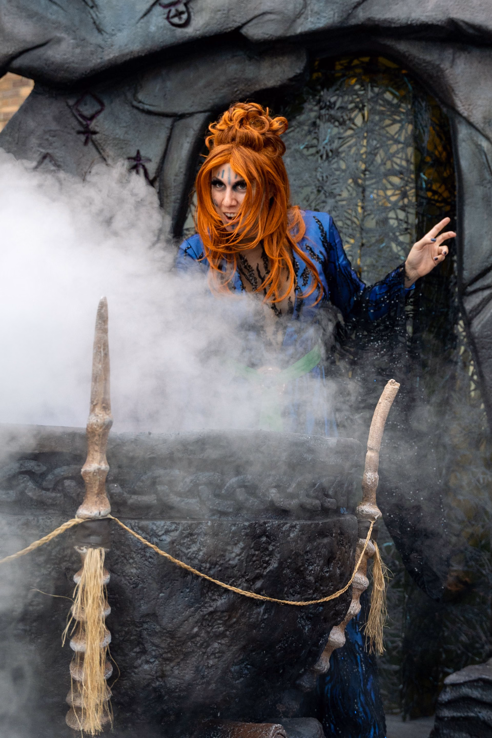 A red-haired woman in a witch costume beside a black cauldron as thick white fog spills over its rim and across the floor