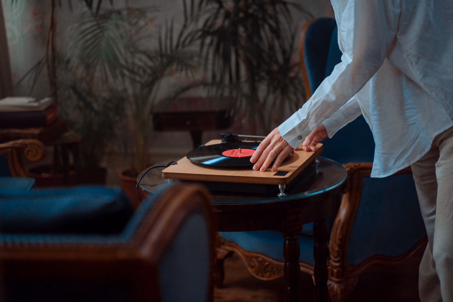 A person in a white shirt setting a record on a wooden turntable on a small round table, in a dim room with a velvet armchair and potted palms