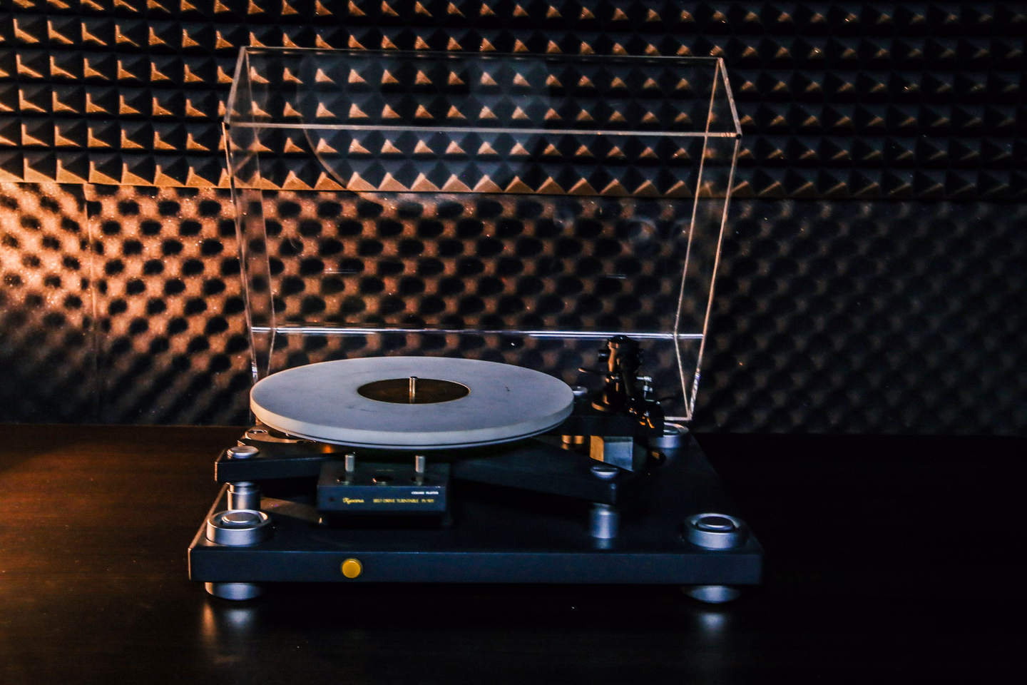 A turntable with a clear dust cover raised, standing on a dark table in front of a wall of black acoustic foam, lit from the side