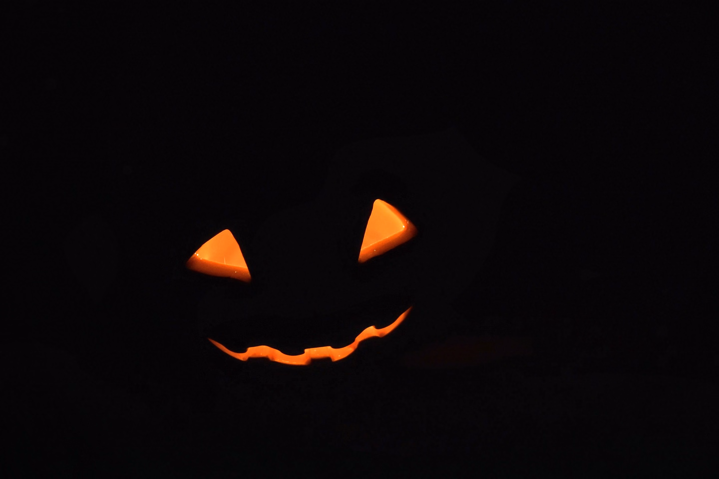 A jack-o'-lantern's triangle eyes and jagged grin glowing orange in total darkness, the pumpkin itself barely visible
