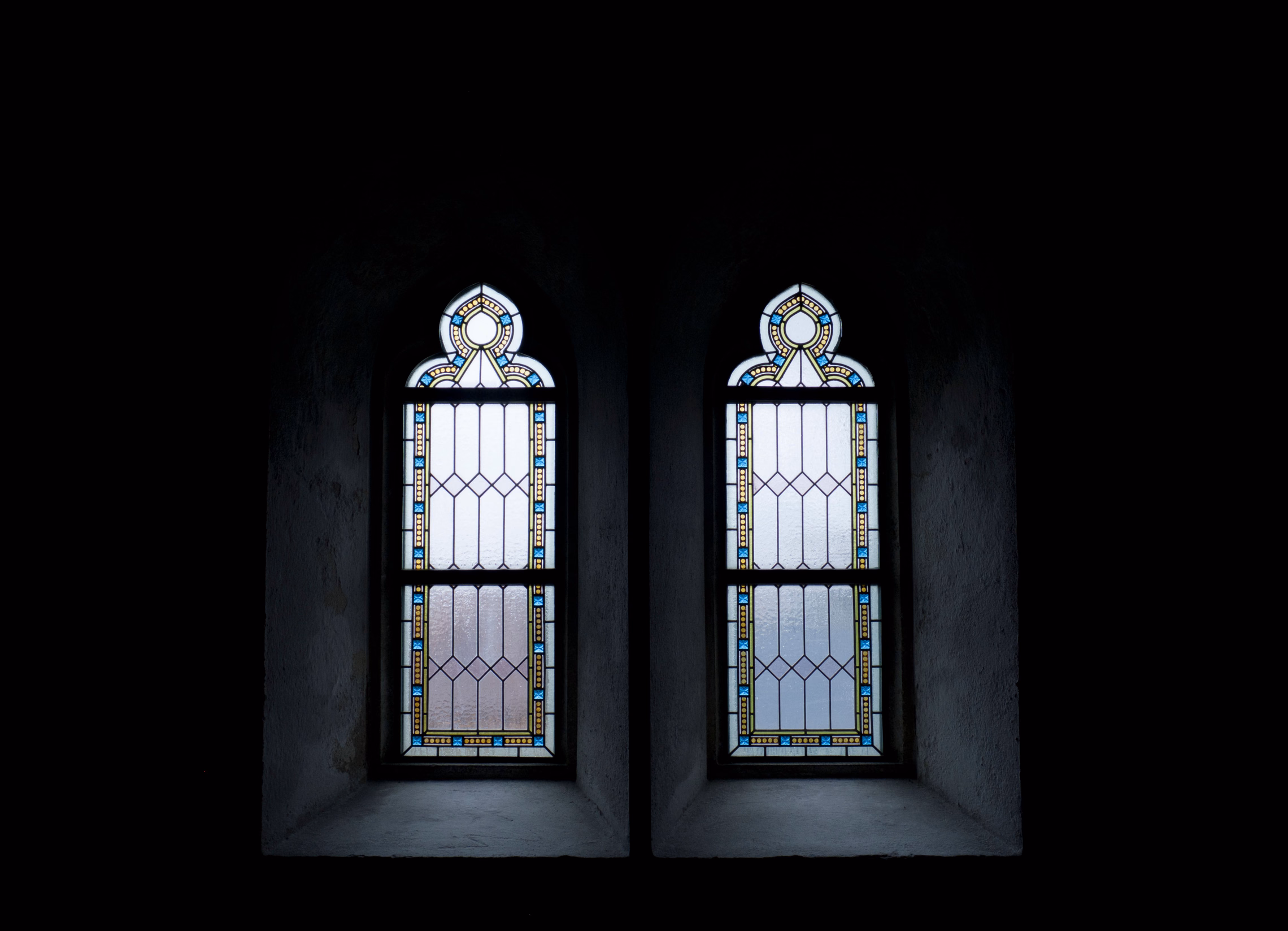 Two tall pointed windows side by side in a dark stone wall, clear leaded panes edged with a thin border of blue and amber glass, the rest of the room in shadow
