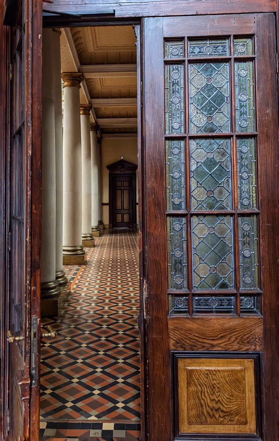 An open wooden door with a tall panel of real leaded glass in small rectangular panes of amber, green and pale glass, looking into a hallway with a patterned tile floor and columns