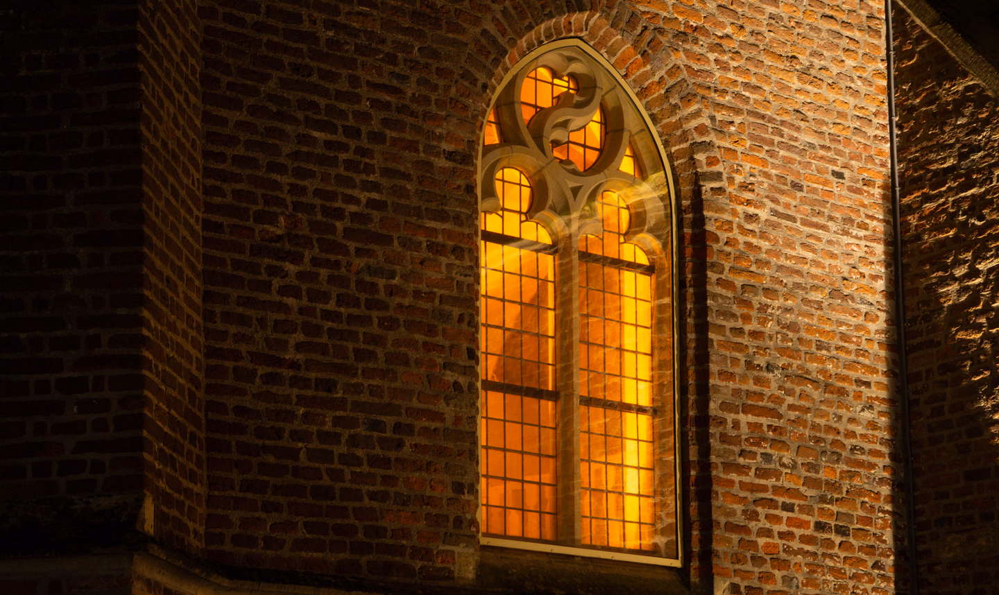 A tall pointed church window in a brick wall at night, glowing solid orange from light inside, with round tracery shapes at the top standing out in black silhouette