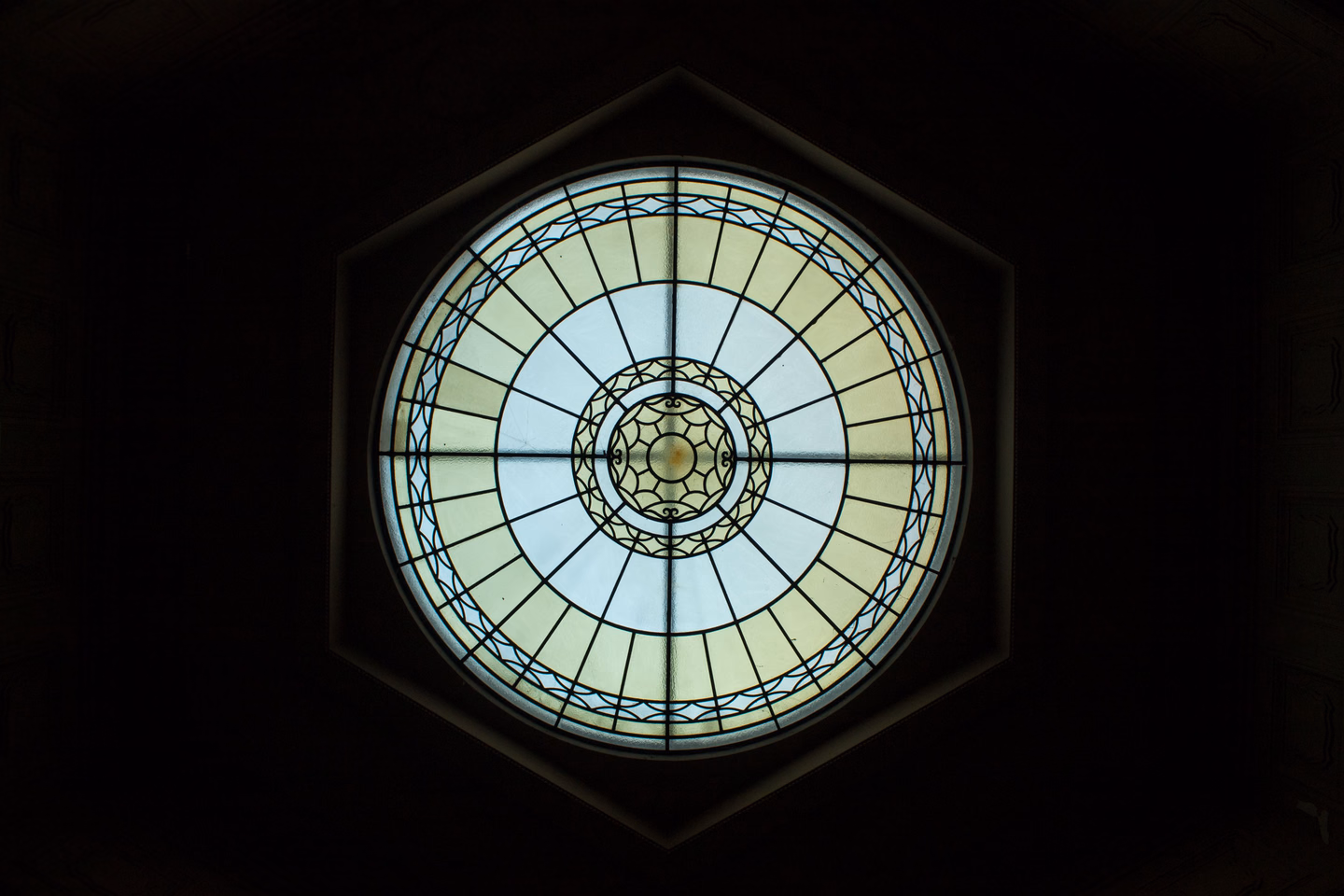 A round window seen from inside a dark building, pale yellow and white glass divided by radiating lead lines around a small patterned center, set in a hexagonal frame