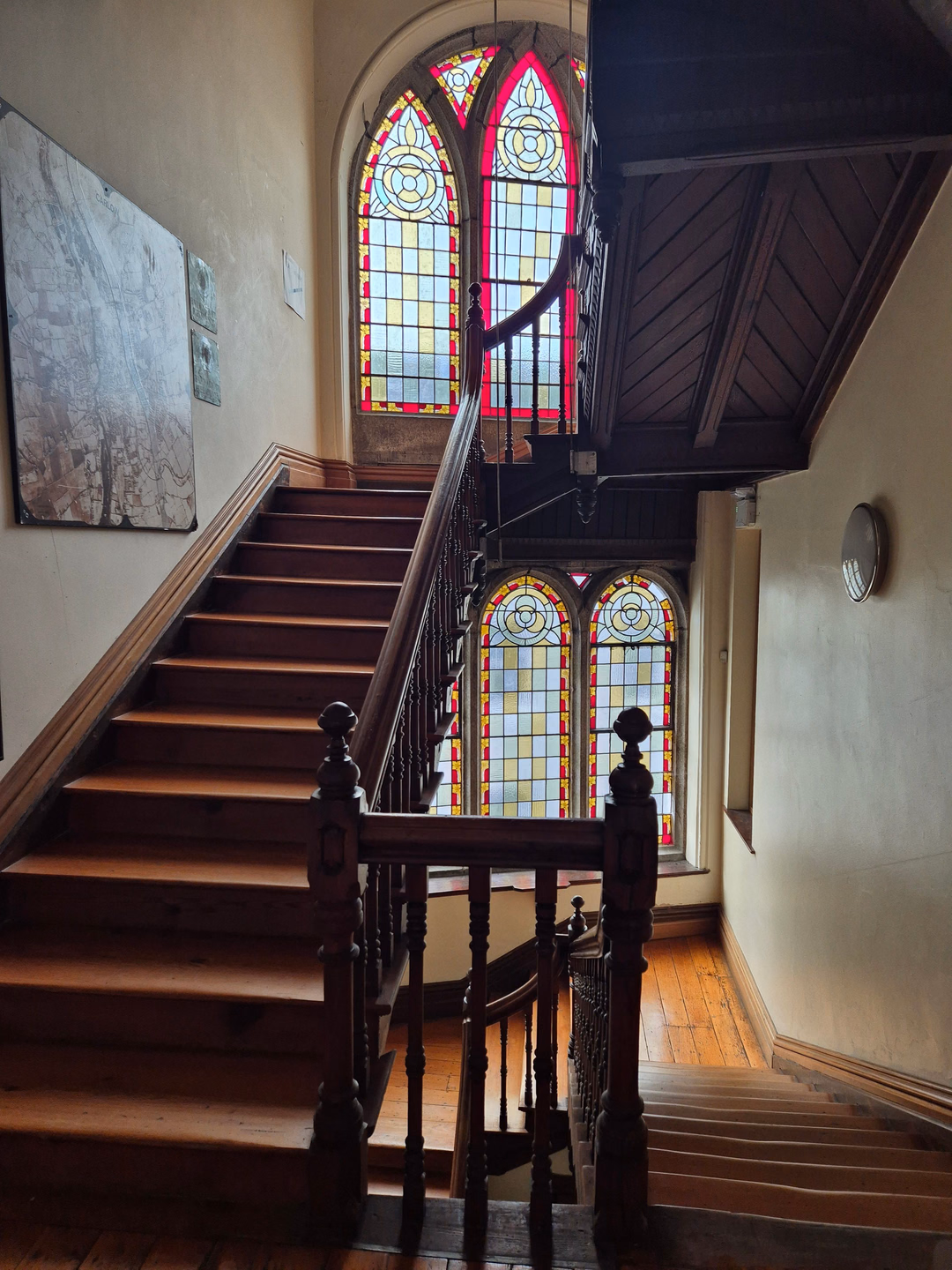 A dark wooden staircase with turned balusters climbing past two pairs of tall arched stained glass windows in red, blue and amber, in an old house with paneled walls