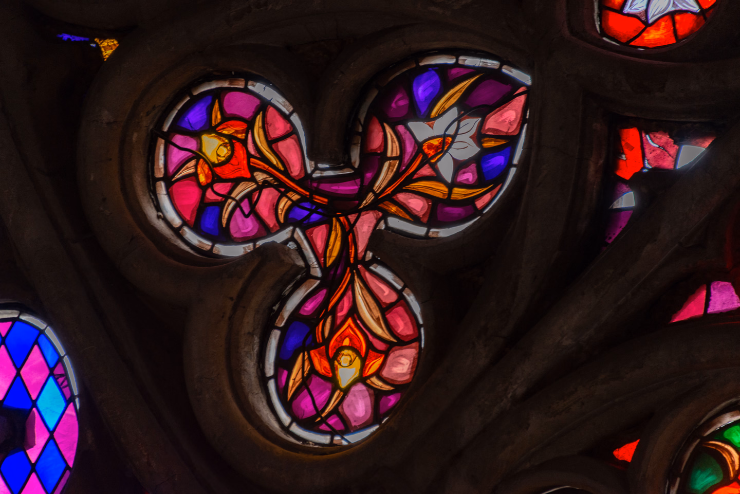 Close view of three round openings in dark stone tracery, filled with glass in deep pink, purple, cobalt blue and amber, with painted lily shapes