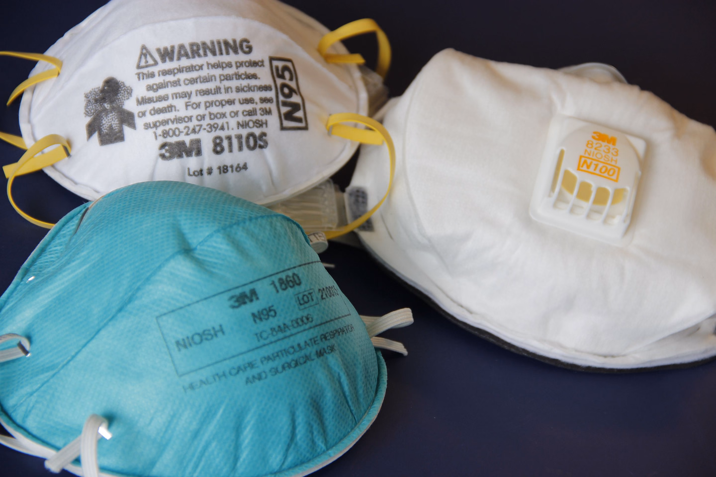 Three NIOSH-approved N95 respirators on a blue surface, one teal, one white with yellow straps and a warning label, one white with an exhalation valve