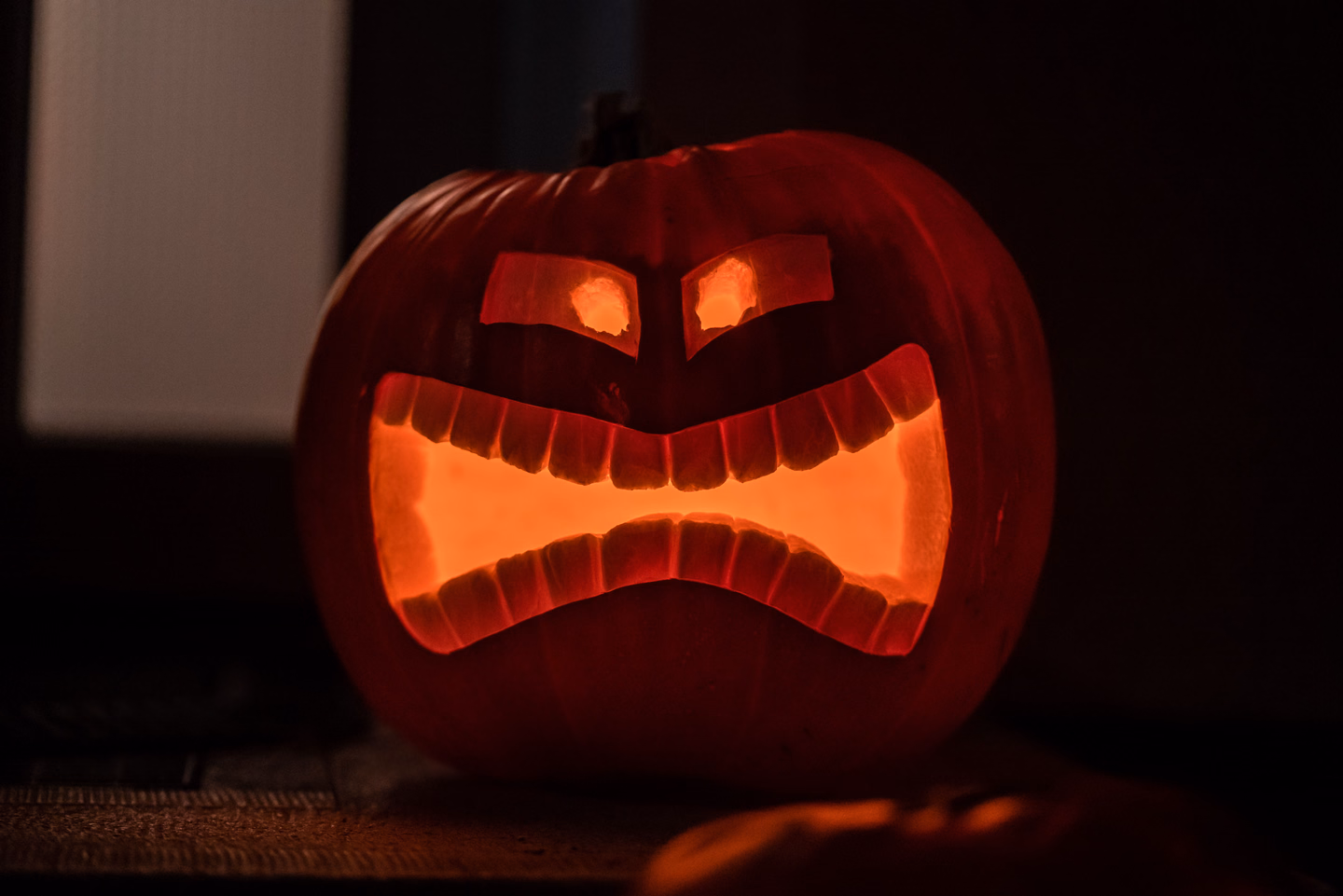 A carved pumpkin with angry slanted eyes and a wide grimace of blocky teeth, lit orange from inside against a dark room
