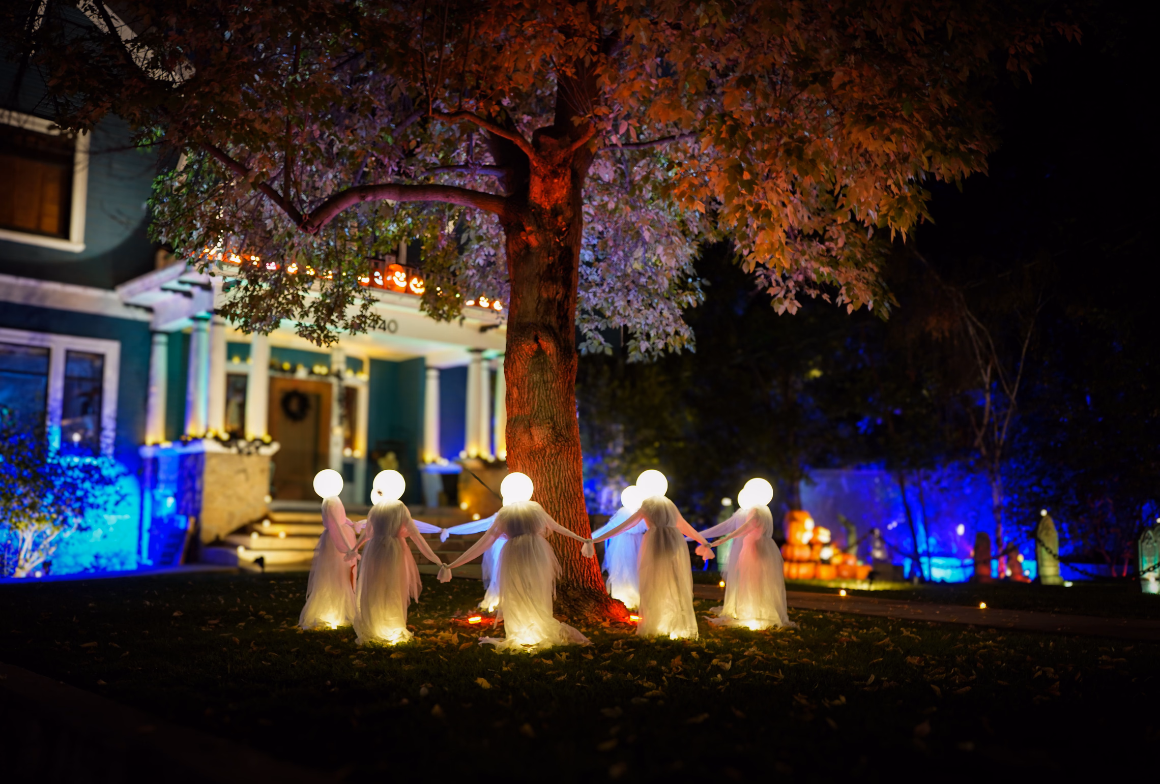 A ring of glowing white ghost figures holding hands on a dark front lawn under a large tree, a porch lit orange and a house washed in blue light behind them