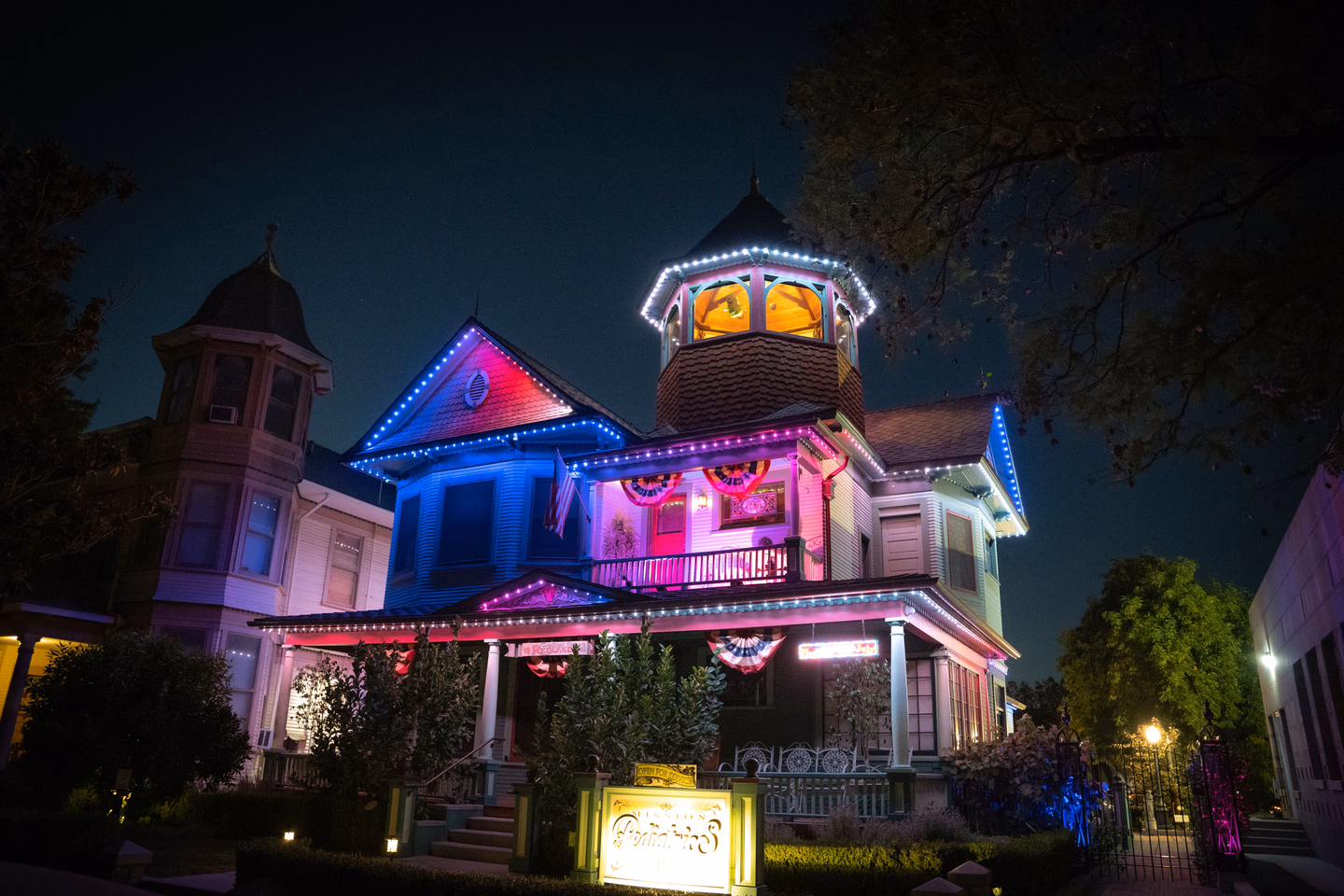 A Victorian house at night washed in saturated pink, blue and purple light, with lit trim along the rooflines and a turret glowing gold