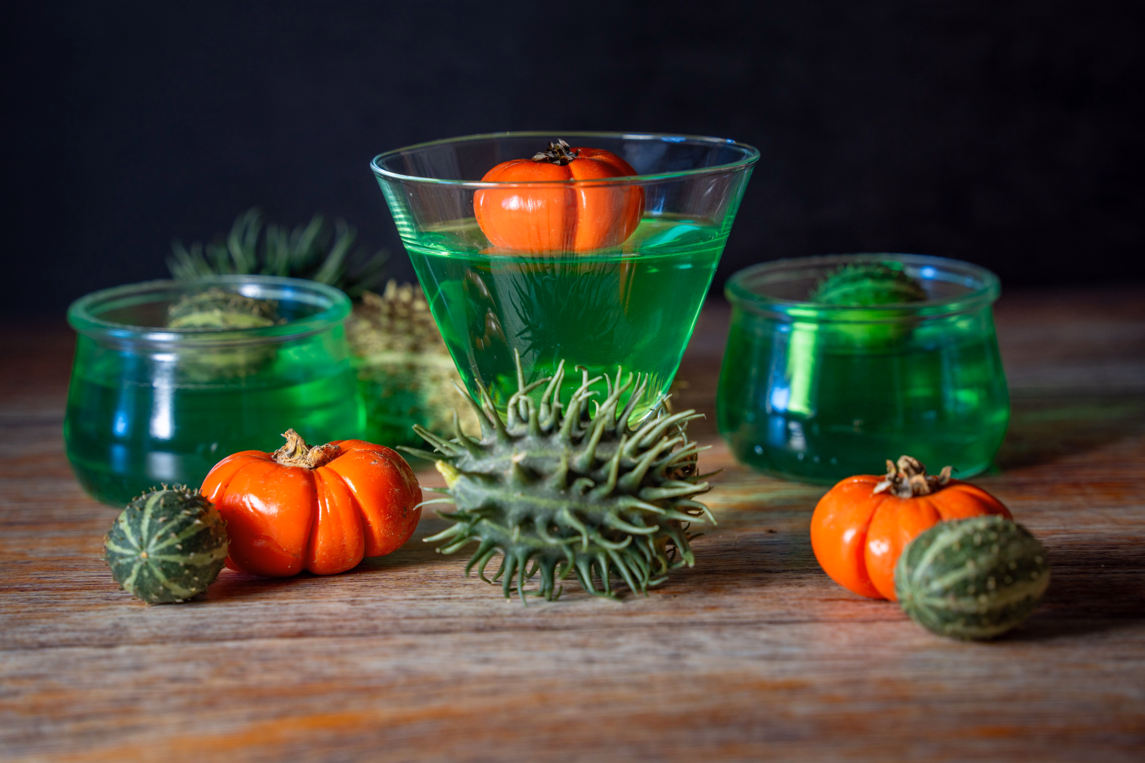 Three glass cups of bright green gelatin on a dark wooden table, a miniature orange pumpkin set into the tallest one, with more mini pumpkins and spiky green gourds around them