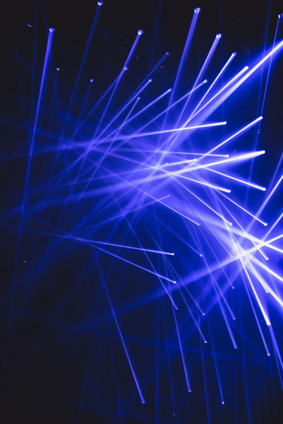 Dozens of blue-violet light beams crossing at different angles against a black background, a white fixture glow at the right