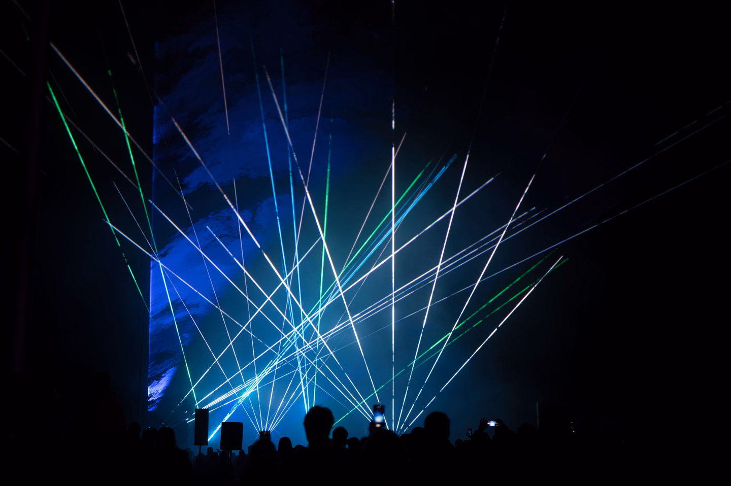 Blue, white and green laser beams crisscrossing high above the silhouetted heads of a concert crowd, a few phones held up to film