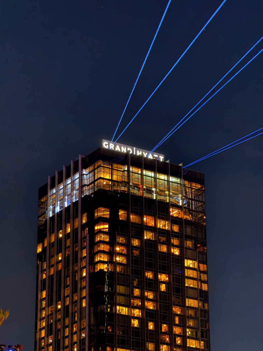 Five blue light beams shooting from the roof of a lit hotel tower up into a dark night sky