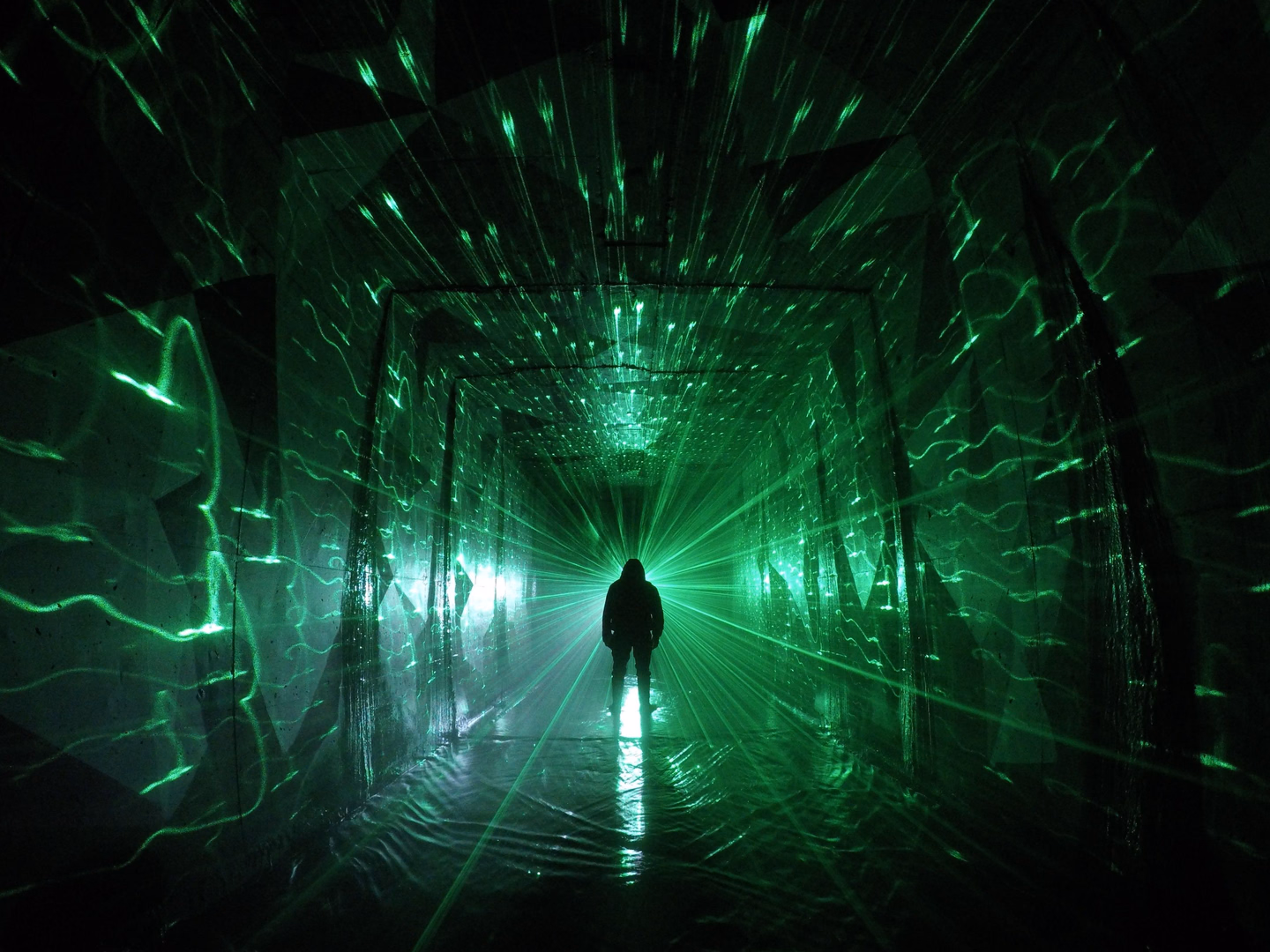 A lone figure silhouetted at the end of a dark corridor filled with scattered green laser light that covers the walls, floor and ceiling