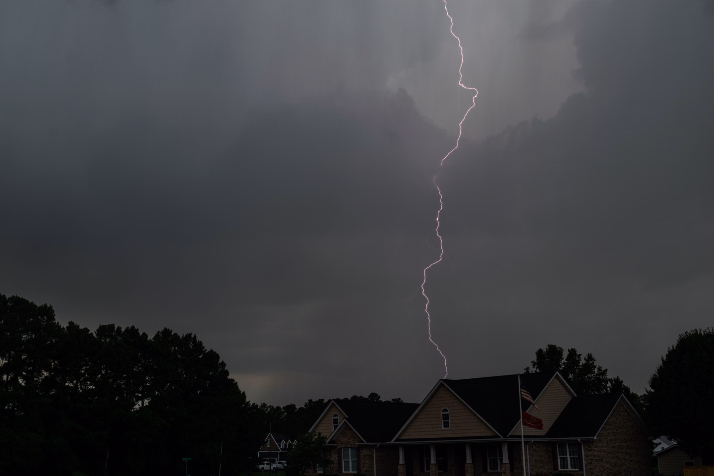 A single jagged lightning bolt dropping from low gray storm clouds toward a two-story house with a flag out front, dark trees on either side