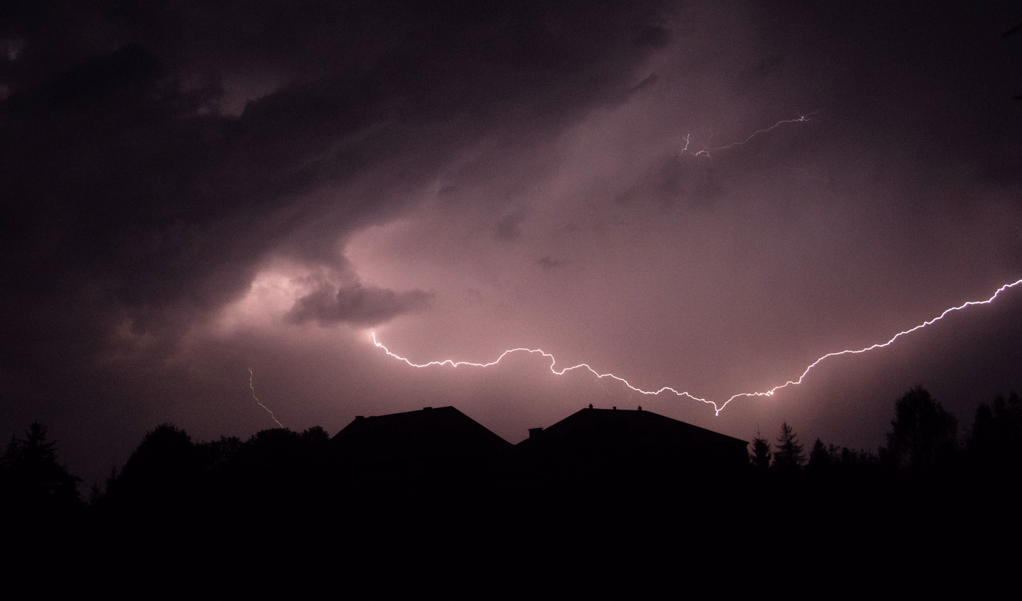 Horizontal lightning streaking across a purple night sky above the dark silhouettes of two house roofs and trees