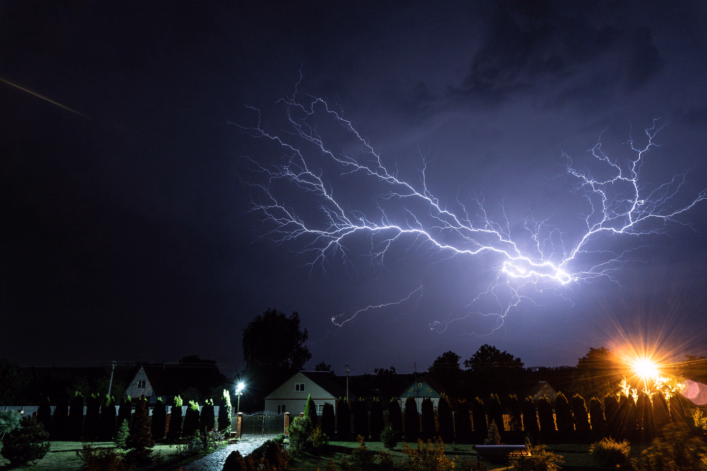 A branching purple-white lightning bolt spreading across a night sky above a row of houses and dark hedges, a streetlight glowing orange at the right