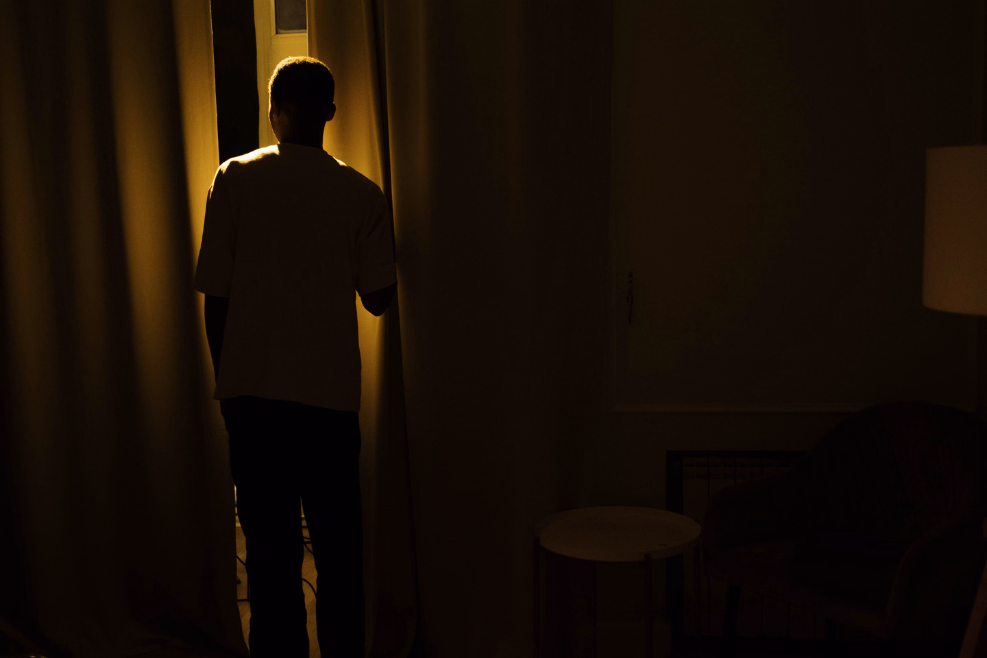 A man seen from behind in a dark room, standing at a gap between long floor-length curtains lit warm yellow from the window