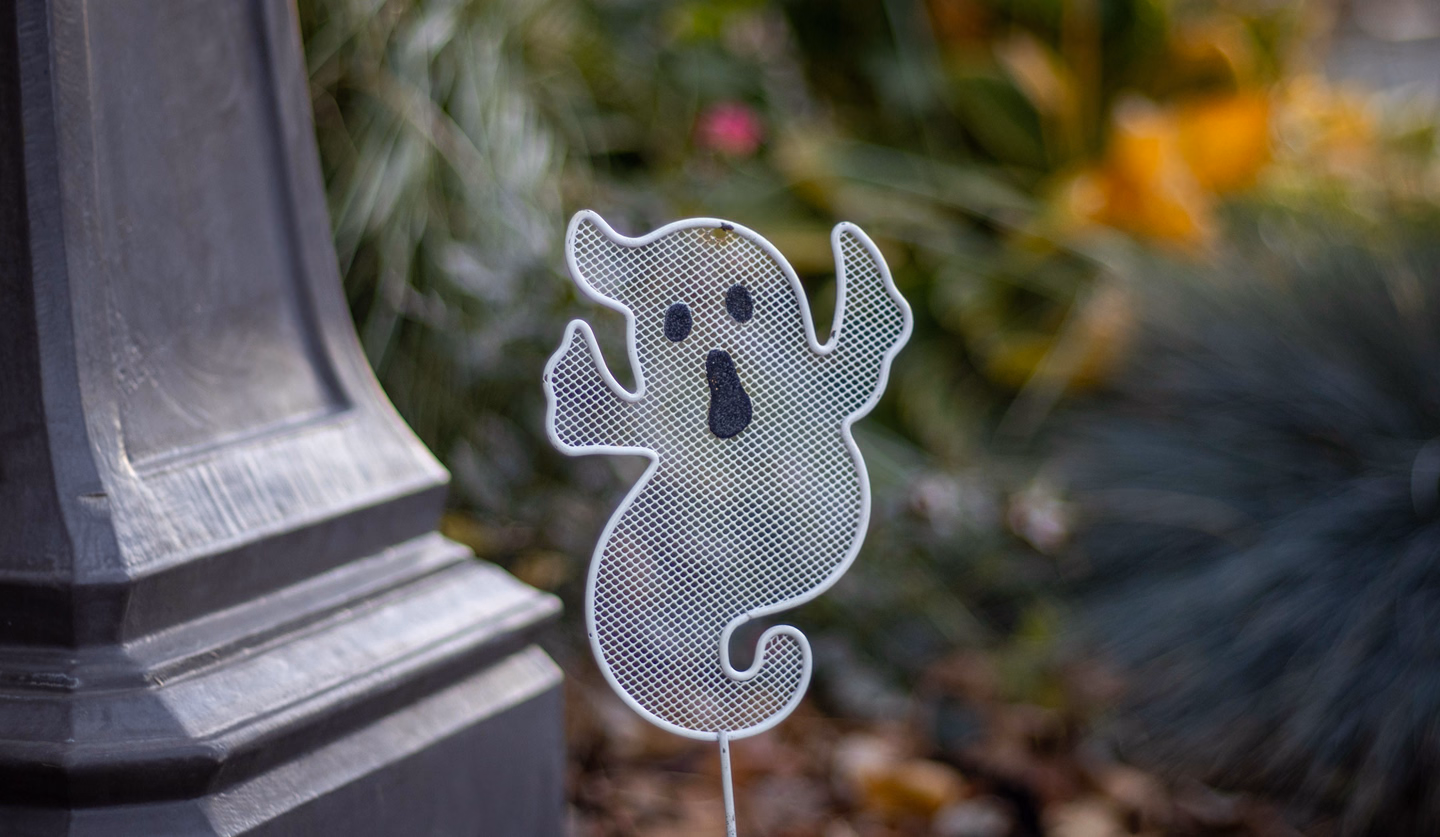 A white wire-mesh ghost yard stake with black eyes and an open mouth, standing in a garden bed beside a dark lamp-post base