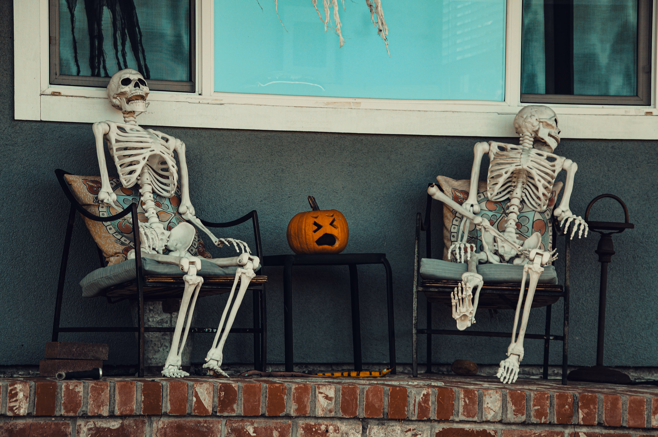 Two life-size plastic skeletons lounging in patio chairs on a brick front porch, a small carved pumpkin on the table between them