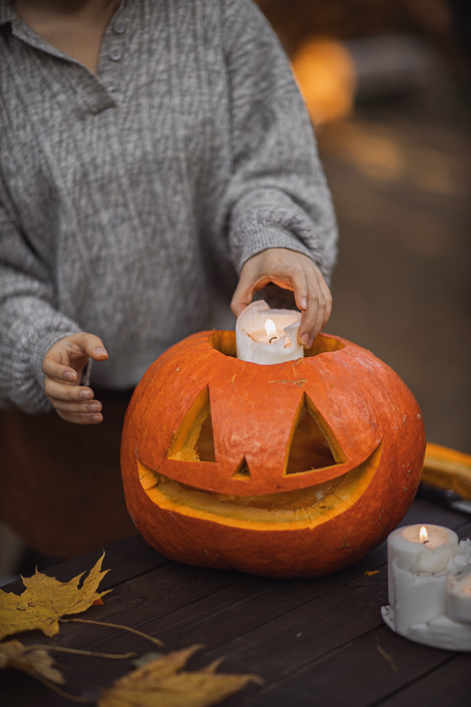 A person in a gray sweater lowering a lit pillar candle into a carved jack-o'-lantern on a wooden table